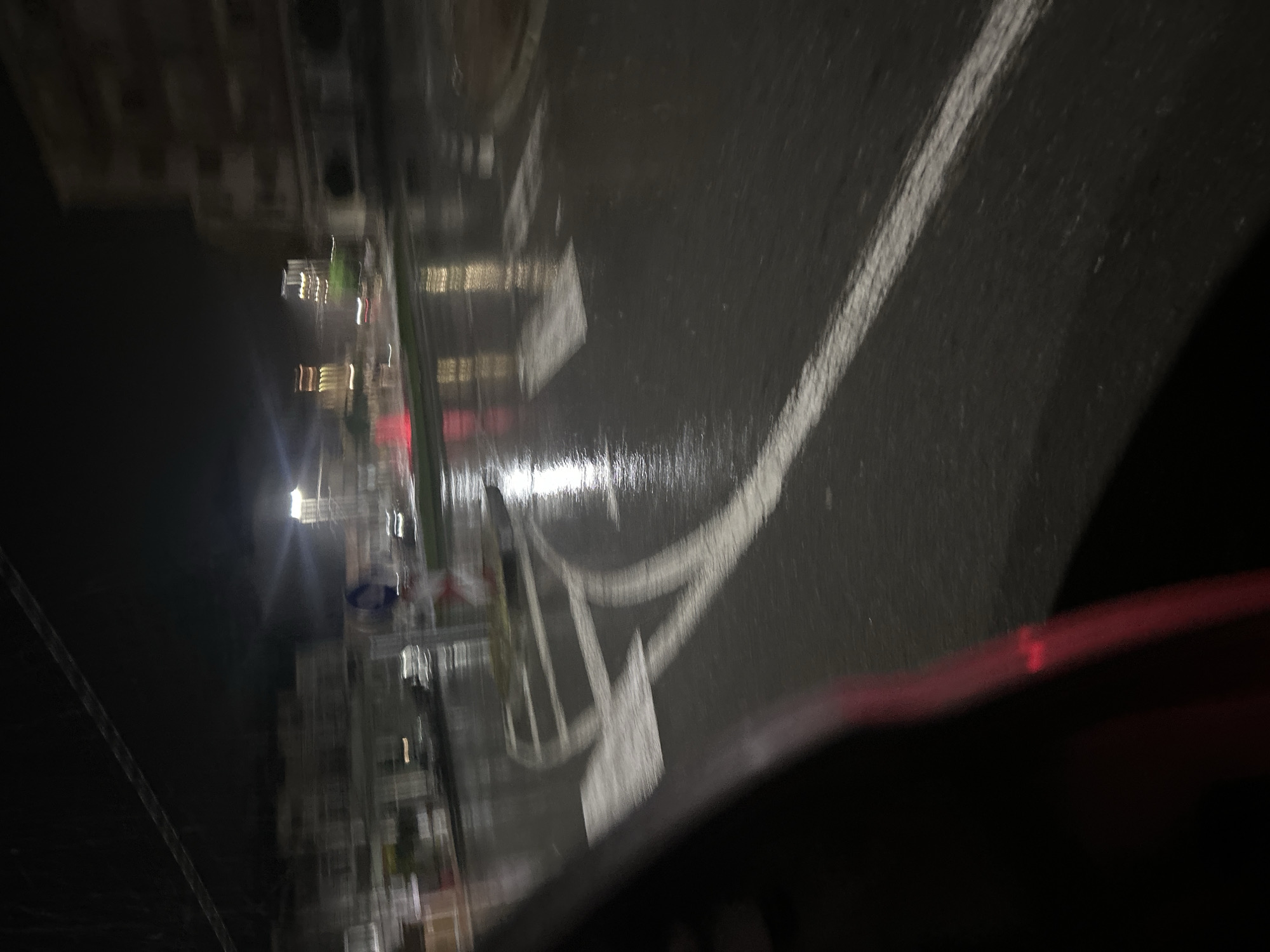 The image depicts an urban street scene at night, likely taken from inside a vehicle. The street appears wet, reflecting ambient light from streetlights and buildings. Road markings, including a crosswalk and lane dividers, are visible. Buildings line the left side of the frame, while the right side is obscured by the vehicle's interior. The image is somewhat blurry, possibly due to movement or low light conditions. The location is unidentified due to the lack of clear, recognizable landmarks or details.