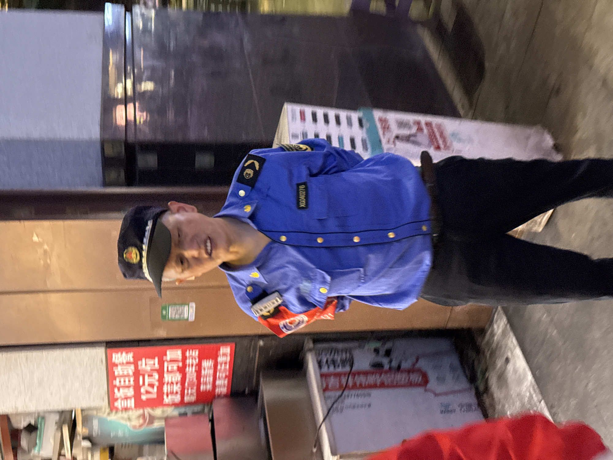 The image shows a man wearing a blue uniform with the label "XG080276" standing on a sidewalk. He also wears a cap with a gold emblem. Behind him, there is a storefront with red signs in Chinese characters. There are also boxes and other objects stacked behind him. The image was likely taken in an urban area, given the signs and the presence of a storefront. The location can be determined to be Yuzhong District, China, based on the writing on the uniform.
