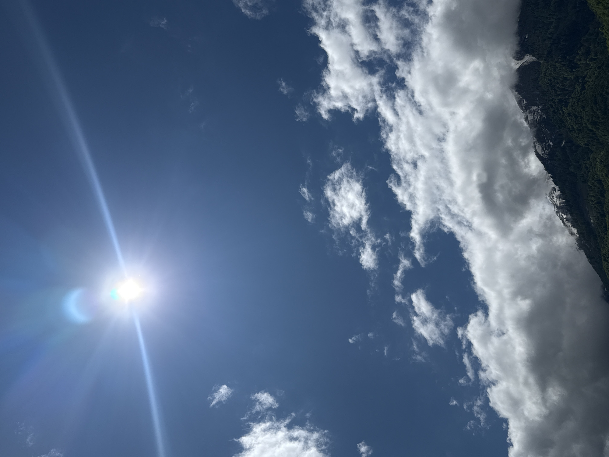 The image shows a bright, daytime sky in Krasnaya Polyana, Russia. The sun is positioned in the lower left, creating a lens flare. There are scattered cumulus clouds in the upper right, some obscuring a green, mountainous ridge. The sky is a clear blue in the areas without clouds.