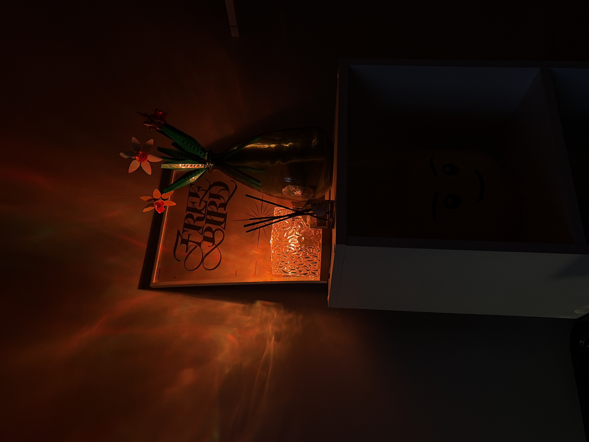The image shows an indoor scene with a shelf or drawer unit in a dimly lit room. On the shelf, there is a glass vase containing LEGO flowers with orange and white petals. Behind the vase is a book with the title "FREE BIRD" in a stylized font. A reed diffuser with black reeds is placed in front of the book, and next to it is a textured glass object that refracts light. The room is illuminated by warm, orange-toned light that casts interesting patterns on the wall. The shelf unit itself appears to be a drawer, with a simple smiley face drawn inside it. The overall mood is calm and artistic, with a focus on decorative elements. The time of day is not specified, but the dim lighting suggests it could be evening or nighttime. There are no visible weather cues. The location context is Leninsk-Kuznetskiy, Russia.