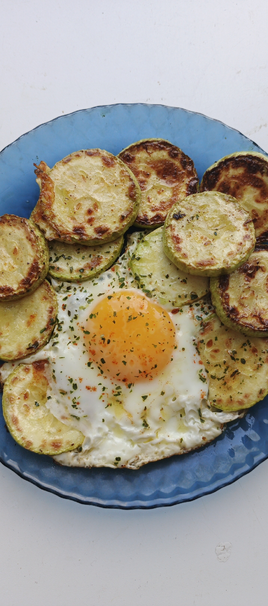A blue plate filled with food sits on a white surface. The food consists of fried zucchini slices surrounding a fried egg. The egg has a bright orange yolk and a cooked white. Green herbs and red pepper flakes are sprinkled on top of the egg and zucchini. The zucchini slices are circular and have a golden-brown color, suggesting they have been fried. The plate is a textured blue glass. The setting appears to be indoors due to the consistent lighting. There are no people visible in the image. There is no location in the city of Chaikovsky, Russia visible in the picture, so I cannot comment on its relevance.