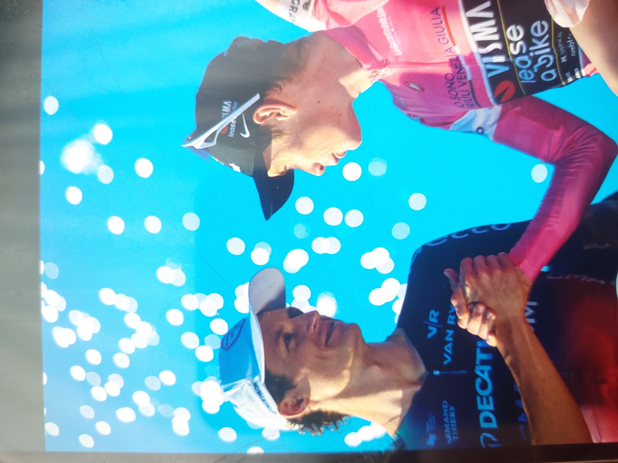 The image shows two cyclists, one wearing a pink jersey with "VISMA Lease a bike" and "La Gazzetta dello Sport FRIULI VENEZIA GIULIA" printed on it, and the other wearing a navy blue jersey with "VAN RY Decathlon" and "ARMAND THIERRY" written on it. They are clasping each other's hands. The background is bright blue with white bokeh-like circles. The location is not specified in the image.