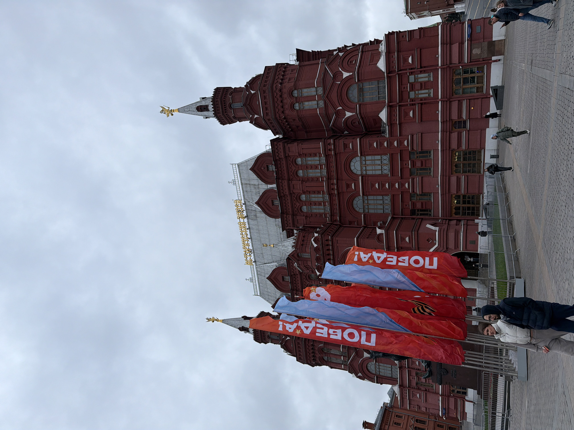 The image displays the exterior of the State Historical Museum, located on Red Square in Moscow, Russia, under an overcast sky. The prominent building is constructed of ornate red brick, characterized by multiple towers, spires, and arched windows with intricate architectural detailing. A golden finial, possibly a two-headed eagle, adorns one of the spires. Along the main roofline, golden letters spell out "ИСТОРИЧЕСКИЙ МУЗЕЙ" (Historical Museum).
In the foreground, several large red and light blue banners are prominently displayed on flagpoles. The red banners feature white text, clearly reading "ПОБЕДА!" (Victory!), often accompanied by an exclamation mark. Some of these red banners also show the black and orange striped St. George ribbon, a symbol associated with Victory Day in Russia.
The ground is a paved area, likely part of a public square or plaza, constructed with cobblestones or similar stone tiles. Several people are visible throughout the scene. In the immediate foreground, two individuals are walking past the banners. Further back, near the museum building and in the distance, other pedestrians are scattered across the plaza. Metal crowd barriers are also visible near the flags. The cloudy, grey sky indicates it is daytime. The presence of the "Victory!" banners and St. George ribbons suggests a celebratory or commemorative event, likely around Victory Day (May 9th).