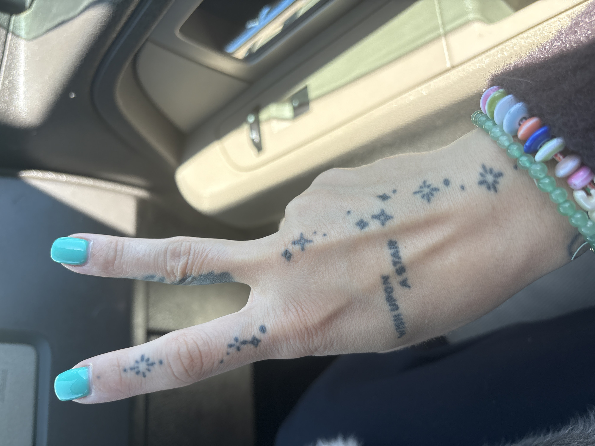 A person's hand, adorned with tattoos and turquoise nail polish, is making a peace sign inside a car. The tattoos are of small stars and the phrase "Wish Upon A Star". The person is wearing colorful beaded bracelets. The interior of the car is beige. The location cannot be determined from the image.