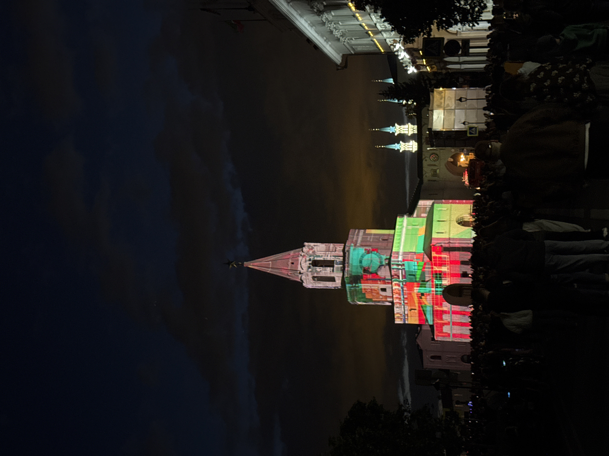 The image shows a night scene in Kazan, Russia. A tower is illuminated with colorful lights. A crowd of people is gathered in the foreground, presumably watching the light show. Other buildings are visible in the background, also lit up. The sky is dark with clouds.