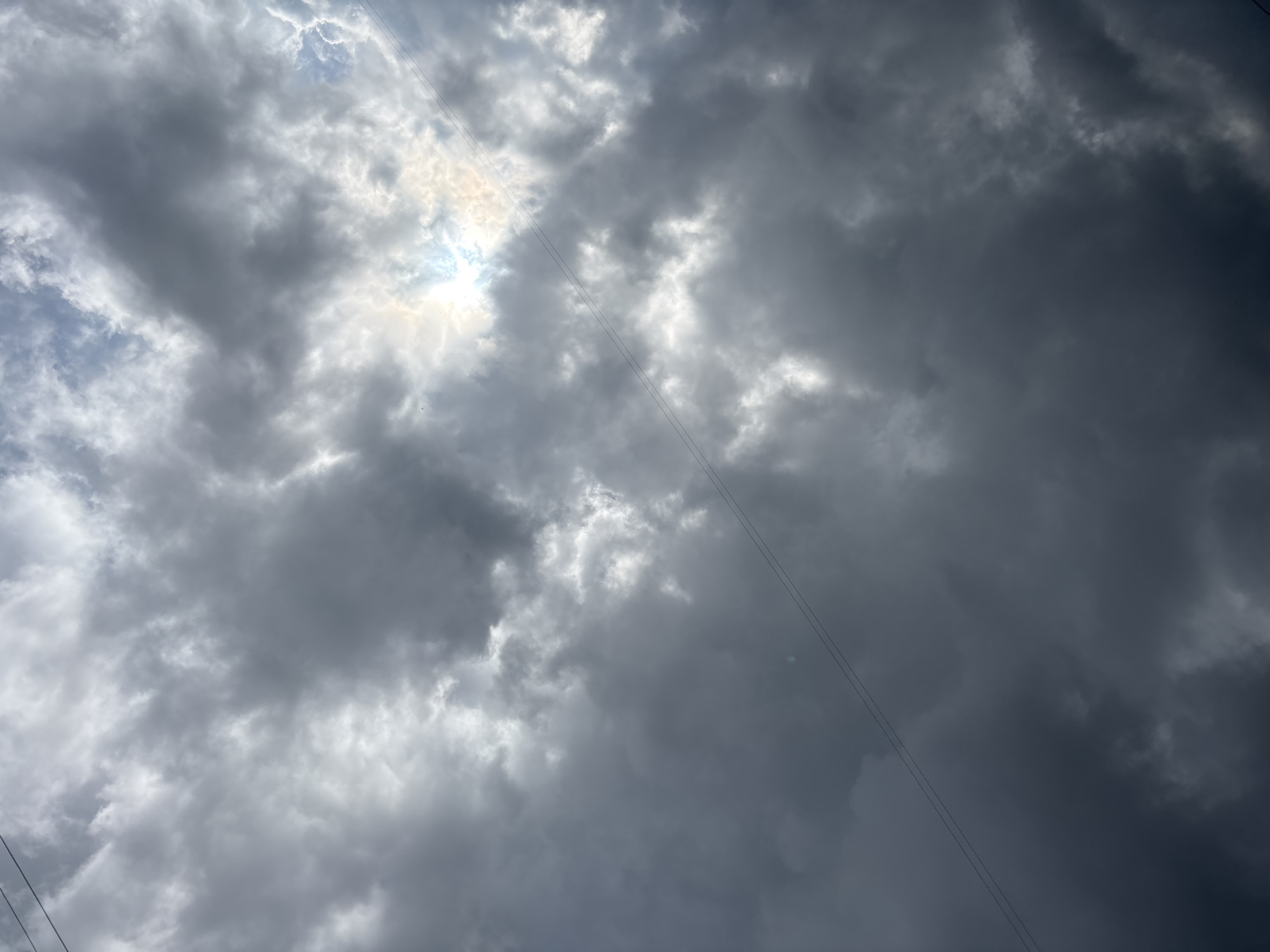The image depicts a cloudy sky over the city of Blagoveshchensk, Russia. The clouds are dark and heavy, suggesting an impending storm or rain. The sun is partially visible, breaking through the clouds in a few places, creating rays of light that illuminate some of the cloud formations. Two thin, dark lines, likely power lines, stretch across the frame diagonally. The overall mood of the image is somber and dramatic due to the heavy cloud cover and the dim lighting.