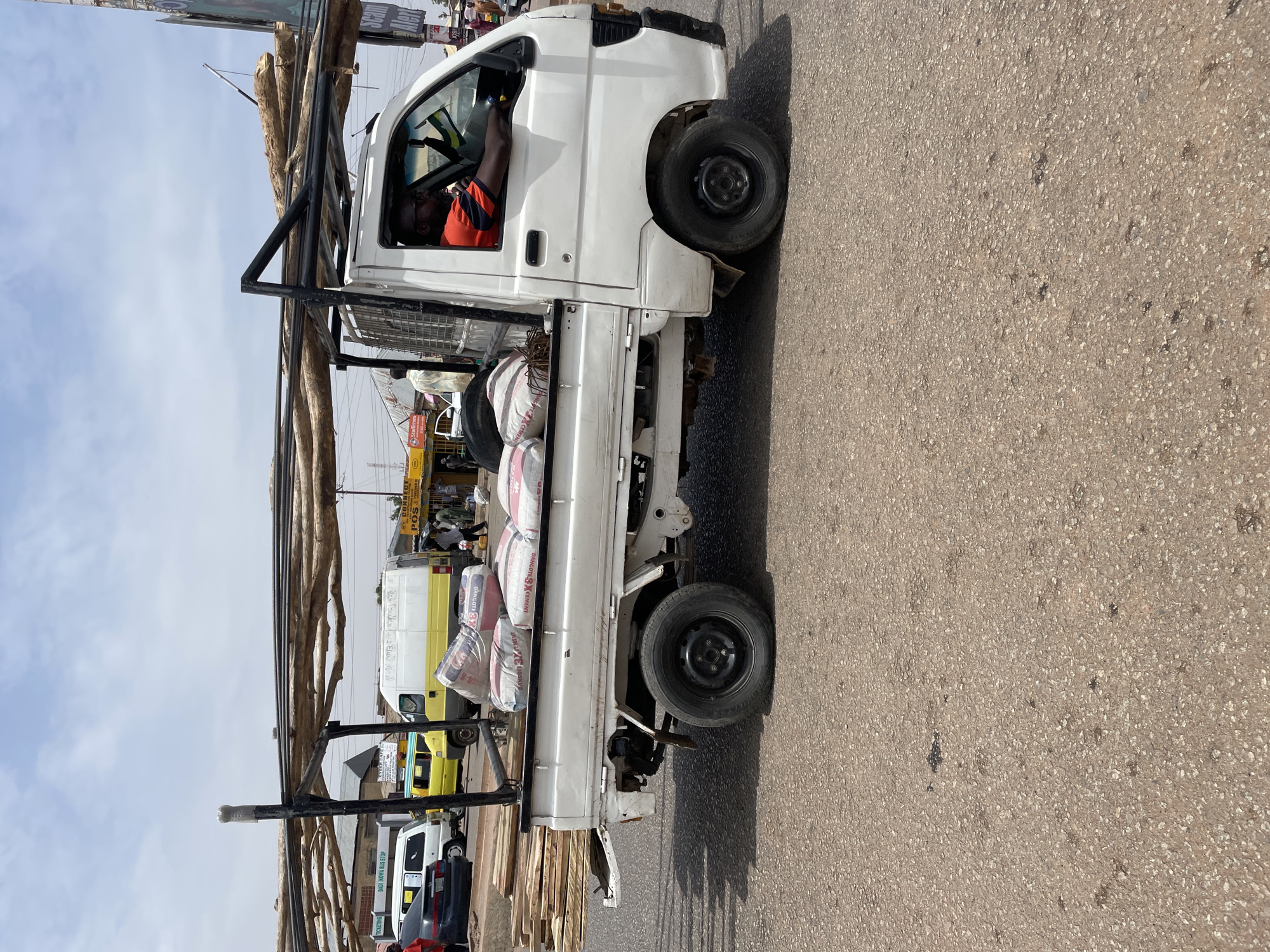 The image shows a white flatbed truck, presumably in Jos, Nigeria, loaded with bags of "Dangote Cement," lumber, and logs secured by a metal frame. The driver is an African man wearing a red and black striped shirt. The truck is parked on an asphalt road. In the background are a yellow and white minivan, other parked vehicles, and a shop with "POS & PHONE" sign. Additional signage includes "DADI IKOWA BUS STOP" and a partial sign "Scam Alert."