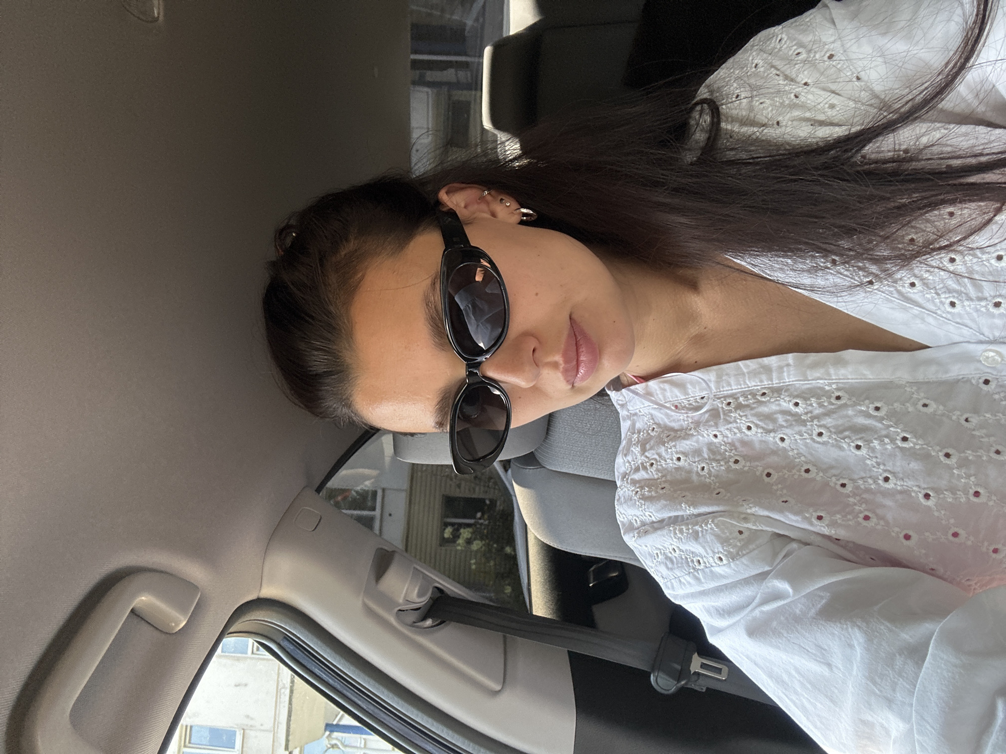 The image shows a young woman with dark hair, wearing sunglasses and a white eyelet shirt, sitting in the passenger seat of a car. She is looking towards the camera, and the interior of the car is visible, including the headrest of the seat in front of her and the ceiling of the car. Outside the car window, buildings and trees can be seen, suggesting an urban or suburban environment. The woman has earrings on both ears. The overall lighting suggests it is daytime.