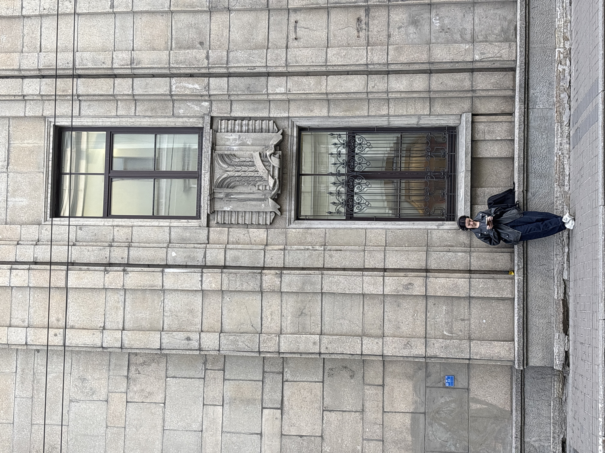 A medium, eye-level, outdoor shot depicts a person standing on a sidewalk against a grey stone building. The building has two prominent windows: one with dark brown gridded panes and the other plain with light reflecting through the panes. There is an ornate stone carving between them. A person wearing a dark jacket, dark pants, white shoes and a hat stands beside the right window. The sidewalk is made of stone tiles. A small, square blue sign is visible on the sidewalk. The architecture is indicative of Saint Petersburg, Russia. The shot appears to be taken from an elevated position.