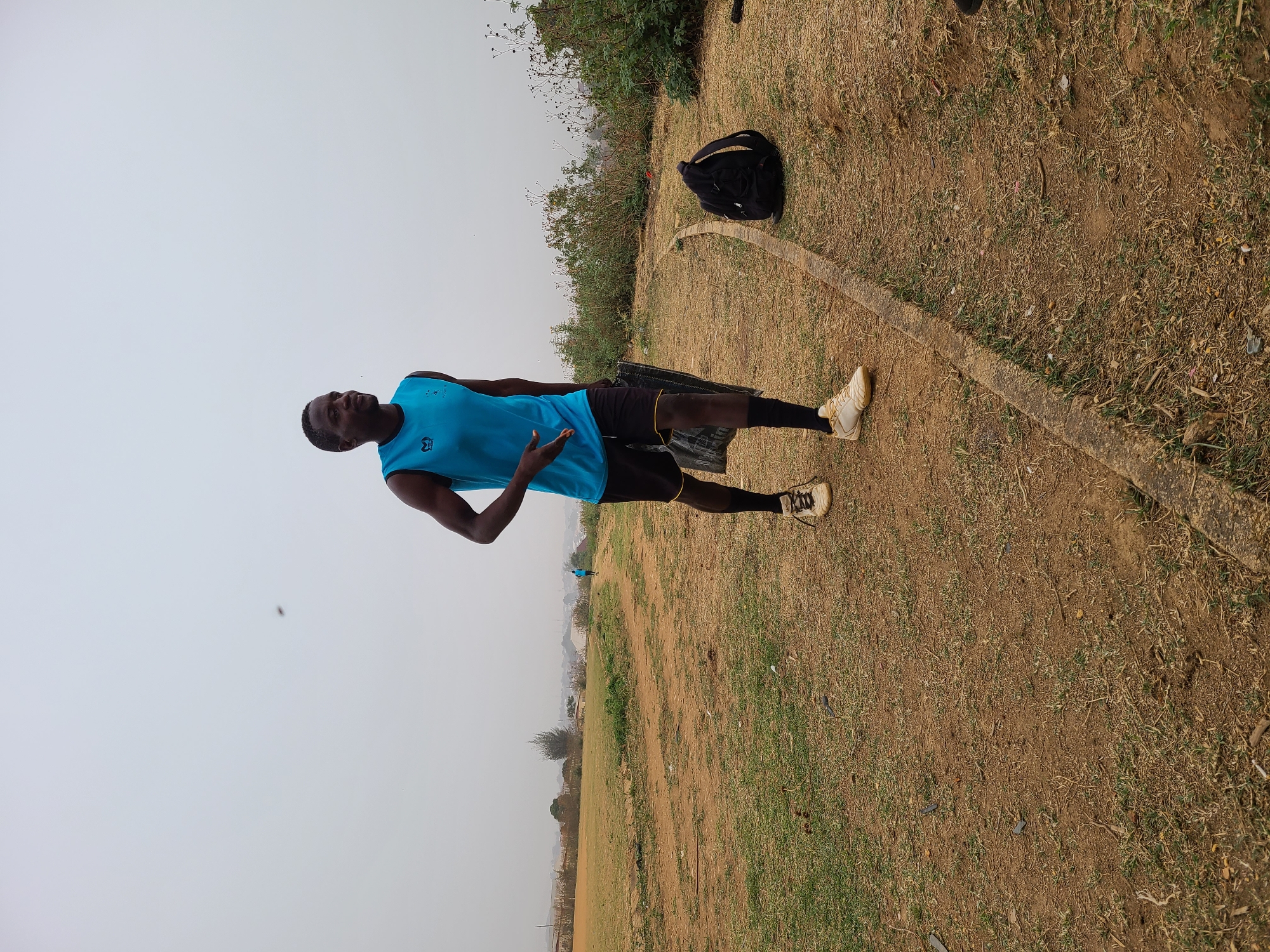 A man in a blue tank top, black shorts, black socks, and tan sneakers stands on a grassy field next to a concrete curb in Barikin Jirga Summa, Nigeria. A black backpack sits nearby on the grass. The sky is overcast. In the distance, the landscape slopes downward.