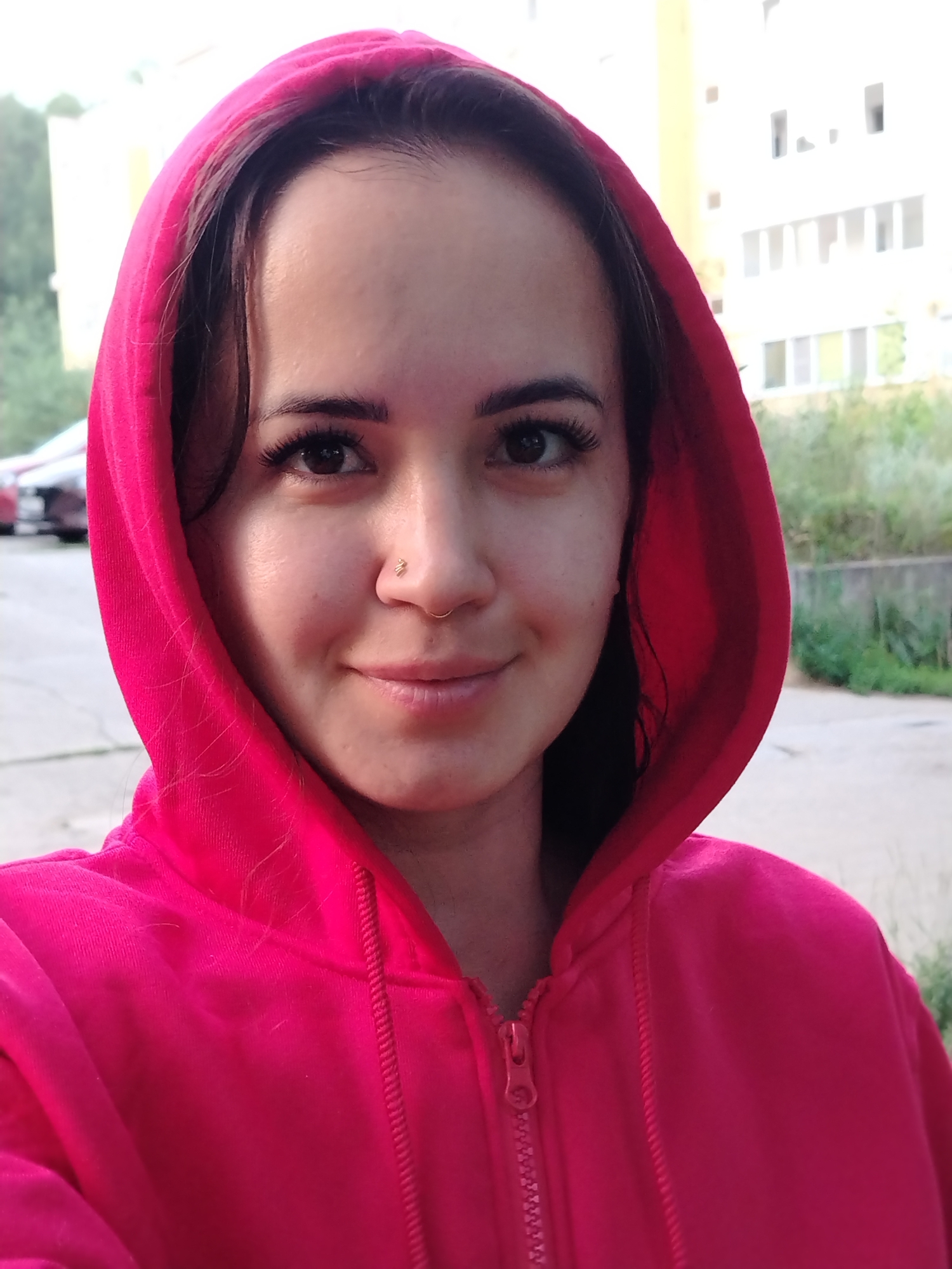 A young woman with brown hair and dark eyes is taking a selfie outdoors. She is wearing a bright pink hooded sweatshirt. She has a small hoop in her nose and a small stud piercing on her right nostril. The background shows a residential building and some greenery. The setting appears to be a street or sidewalk in a city, possibly Noviy, Russia.