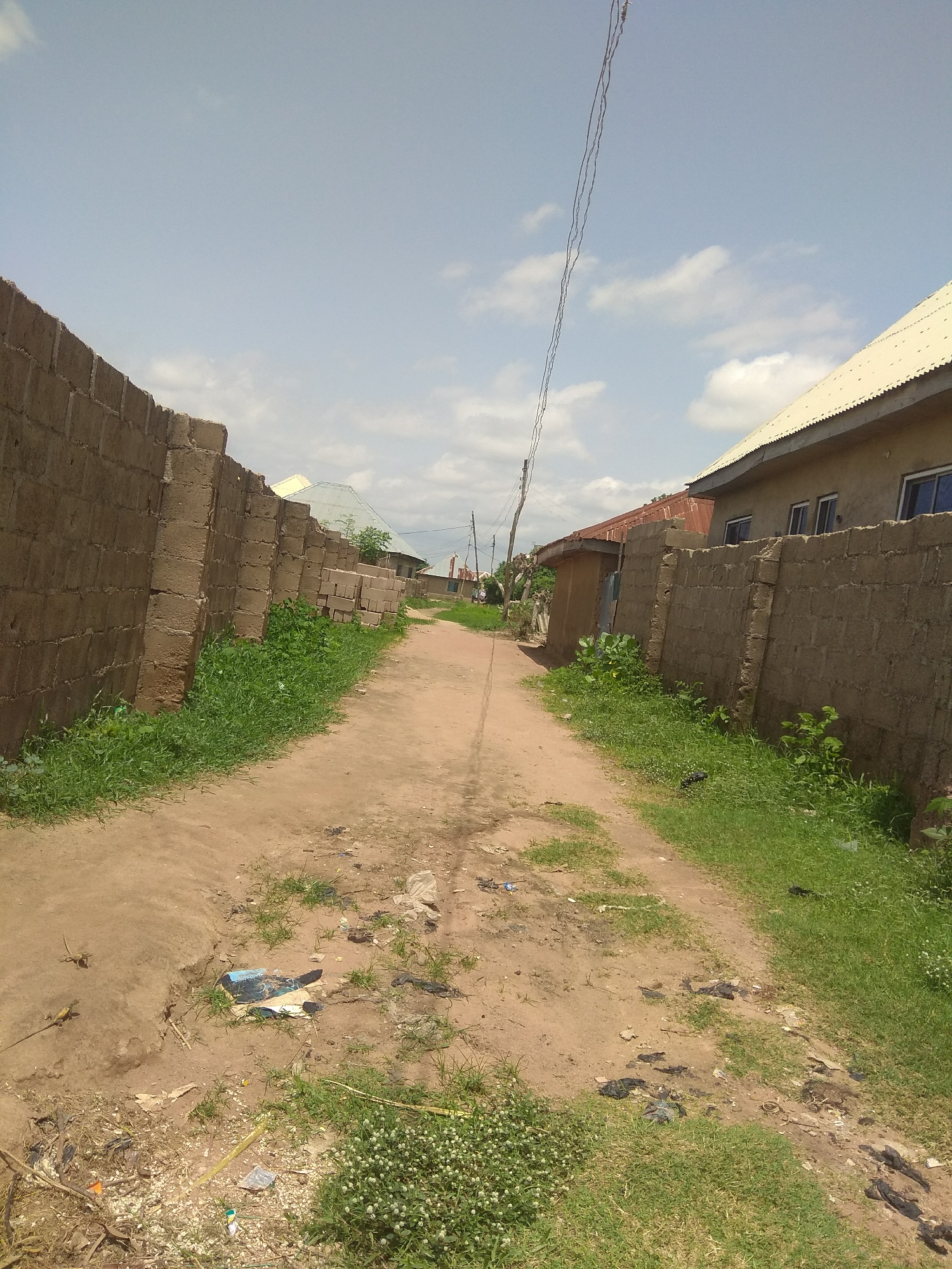 The image depicts a dirt road in Jalingo, Nigeria, lined by stone block walls on either side. The road is unpaved and shows signs of wear, with some loose soil and debris scattered along its surface. To the left, a tall, rough-hewn stone wall stretches into the distance, overgrown with grass at its base. On the right, a similar stone wall borders a residential building with a light-colored corrugated metal roof and several windows. Further down the road, more buildings with corrugated roofs are visible, some with wooden fences.
The sky is partly cloudy, suggesting it is daytime, likely late morning or early afternoon. The weather appears warm and sunny. The scene is one of everyday life in a residential area, with no specific activity or event clearly taking place. The environment seems somewhat unkempt, with litter visible on the road and grass growing wild in some areas. The overall impression is of a typical, modest urban or semi-urban setting in Nigeria. There is no visible text in the image.