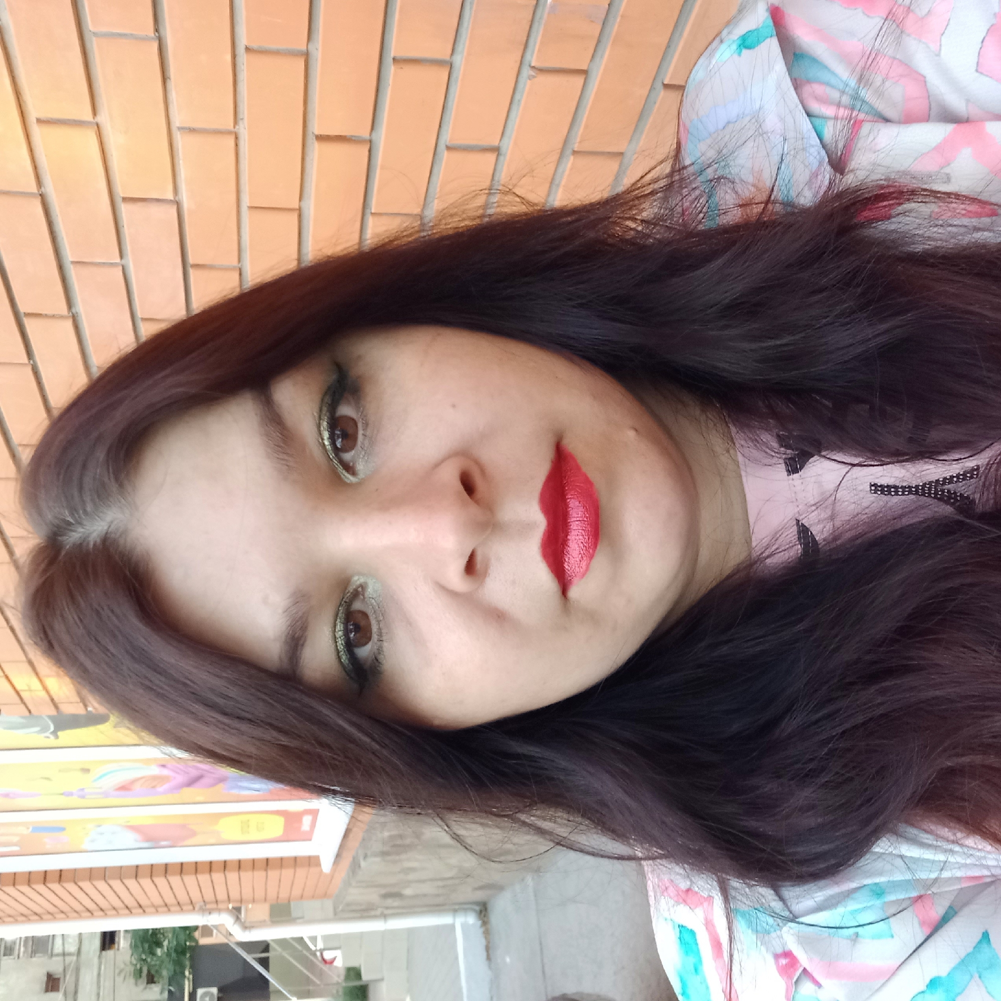 The image is a headshot of a woman with long, dark hair and light skin. She is wearing red lipstick and green eyeshadow. She is standing in front of a brick wall. To her left, there is a building with a window and a sign. The location is the city of Kropyvnytskyi, Ukraine.