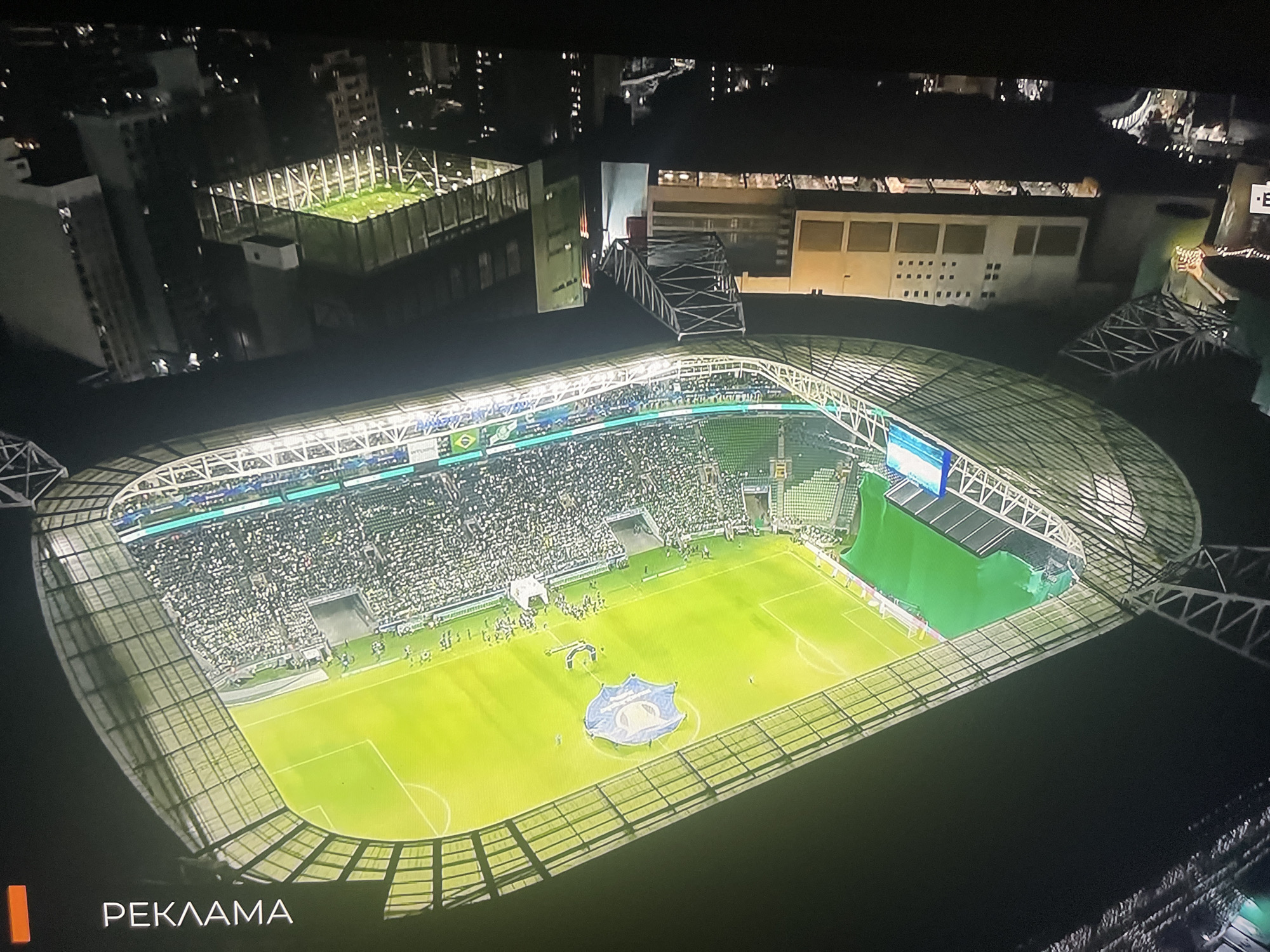 This is an aerial night view of a brightly lit football stadium, likely taken from a drone or helicopter. The stadium is surrounded by a cityscape with numerous buildings visible, some with illuminated windows. The field is green and appears to be in use or recently used, with a large crest or logo laid out on the grass near the center. Spectators are seated in the stands, which are partially covered by a large, latticed roof structure. There are several access points or tunnels visible leading from the stands to the field. In the background, separate from the main stadium, is a smaller, enclosed green space with lighting, possibly a training area. On the bottom left of the image, a text overlay reads "РЕКЛАМА" which translates to "ADVERTISING" in English, indicating this is likely a broadcast or recording. The location is stated as Ruse, Bulgaria.