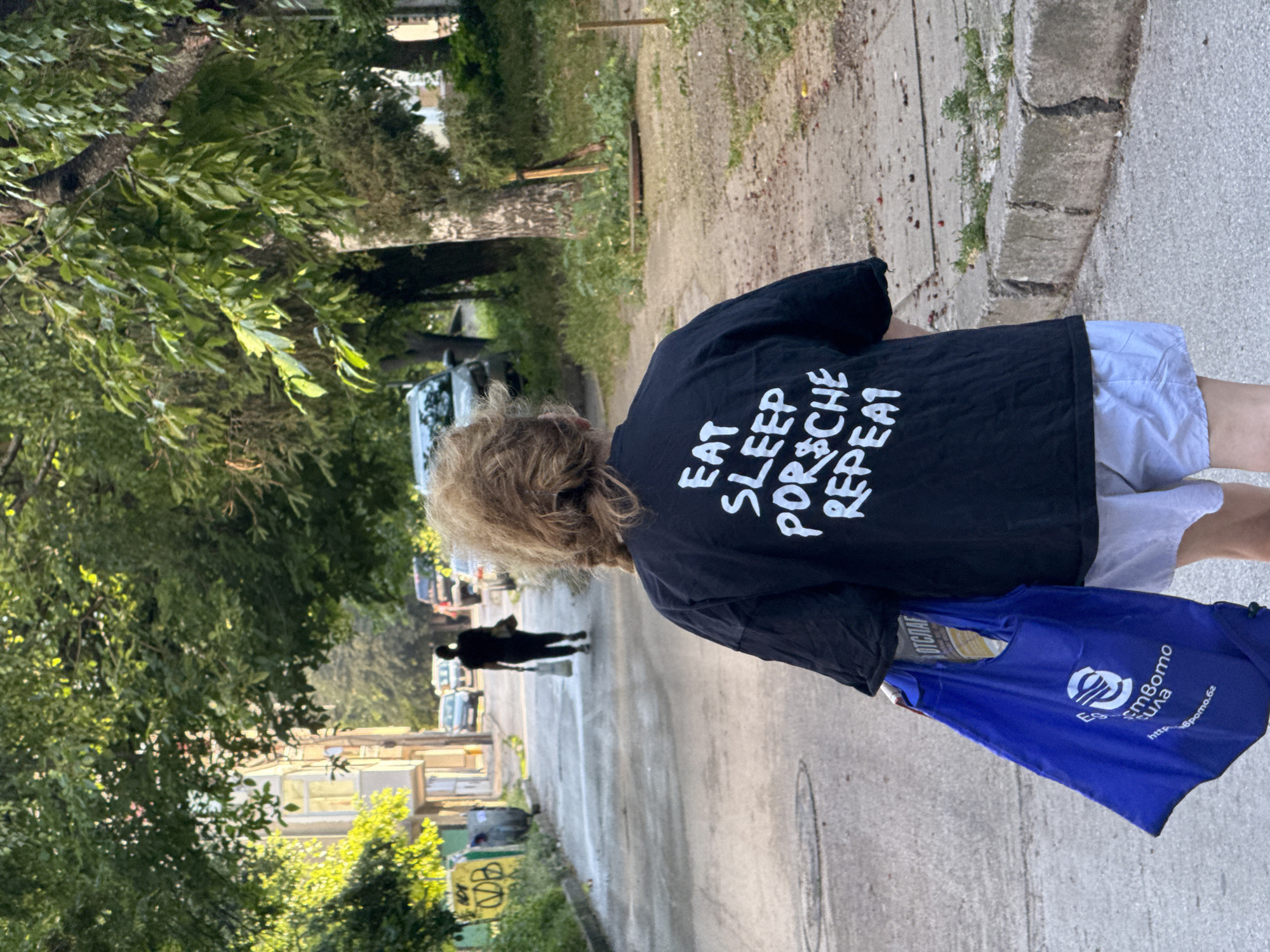 In Pleven, Bulgaria, on a street lined with trees and buildings, a person walks away from the camera. The person wears a black t-shirt with white lettering that reads "EAT SLEEP PORSCHE REPEAT" and light-colored shorts. They are carrying a blue reusable shopping bag in their left hand, which has white text and a circular logo on it. In the background, other people are walking, and cars are parked and moving along the street. The scene appears to be a residential or semi-commercial area.