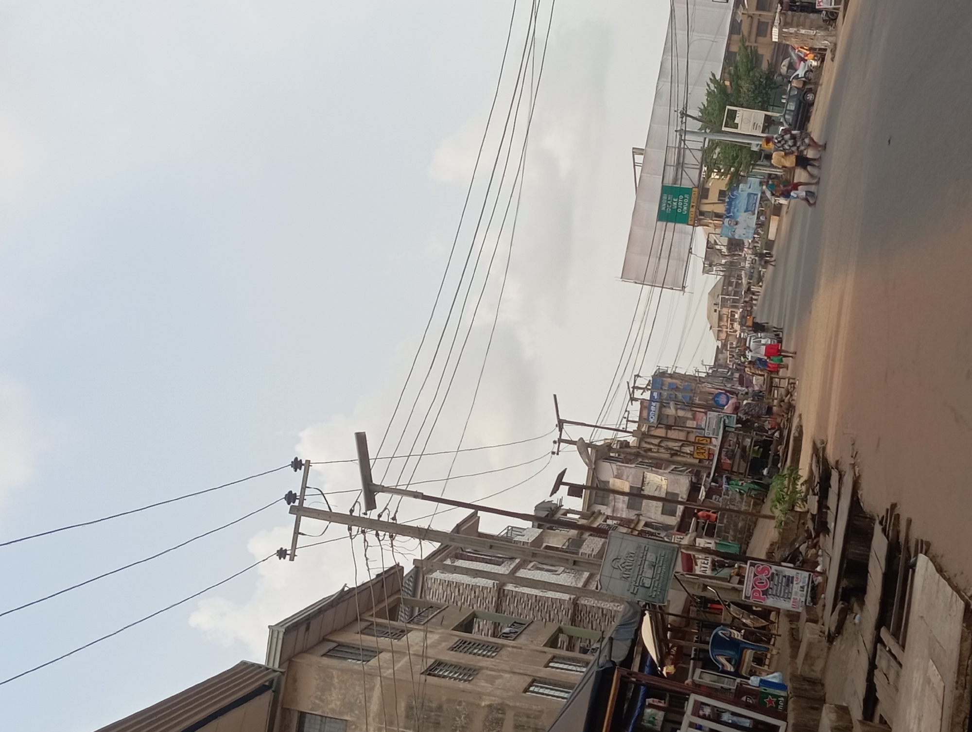 This image, originally tilted, depicts a vibrant street scene during the daytime in Nkpor, Nigeria. The sky is bright with scattered clouds.
The central focus is a bustling commercial street, lined with multi-story buildings that appear aged, constructed from concrete, and often feature barred windows for security. Utility poles with numerous interwoven overhead power lines crisscross the frame, indicating a dense urban infrastructure. The road itself appears unpaved or poorly maintained, with visible cracks and uneven surfaces suggesting wear and tear.
Several people are present, mostly pedestrians, walking along the roadside or crossing the street. In the mid-ground, a group of individuals, one wearing a yellow top, moves across the road. Other people are seen near various stalls and shops that line the street, engaging in daily activities such as commuting or commerce.
Notable objects include a few parked or slow-moving vehicles in the distance, and numerous commercial signs and billboards. A prominent green overhead sign in the mid-ground serves as a key location cue, displaying "ANWULI JUNCTION 1KM", "AWKA, ONITSHA EXPRESS", and "UMUOJI", with smaller text "INVOICE IBADAN" on its side. Other visible text includes "AKUGO" on a building sign, accompanied by what appears to be Arabic script, and "POS" on a smaller sign, indicating Point of Sale services. Various temporary stalls and displays of merchandise, including a blue plastic chair, are arranged along the street, underscoring the commercial nature of the area. The scene conveys a sense of everyday life and activity in a busy Nigerian town.