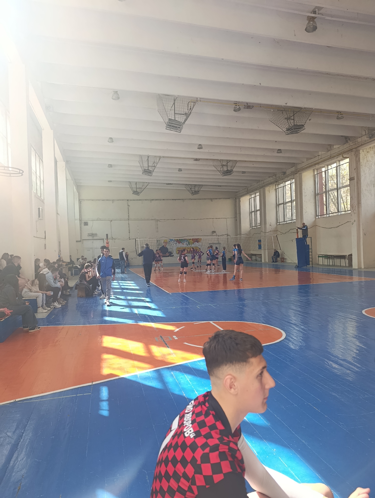 The image shows a volleyball game taking place in what appears to be a school gymnasium. The walls and ceiling are painted white, with metal cage-like structures hanging from the ceiling. Natural light streams through the windows, casting shadows on the orange and blue painted basketball court.
A volleyball net stretches across the middle of the court, and two teams of young women are playing. There's a man standing on a platform, officiating the game. Spectators sit on benches along the side of the court.
In the foreground, a young man is seated, facing towards the game. He is wearing a red and black checkered shirt with white sleeves. The inscription on the shirt is Russian. The location is likely in Silistra, Bulgaria, as it appears to be the city referenced by the Russian inscription.