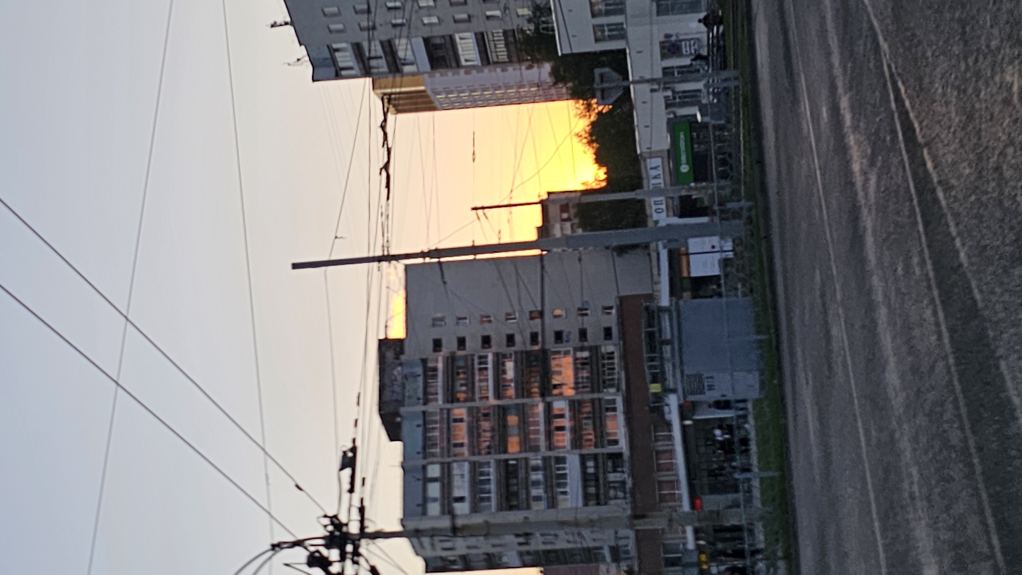 This image depicts a street scene in Yekaterinburg, Russia, during sunset. The sky is a pale gradient from light blue to warm orange, indicating the setting sun. Dominating the mid-ground are several multi-story apartment buildings. The building on the left is gray and appears to have many windows reflecting the sunset's light. To its right, a slightly taller, light-colored building with golden hues is visible, its upper floors obscured by trees. A modern, white building with many windows is situated on the far right. Overhead, a complex network of power lines and utility poles crisscrosses the frame, casting shadows. In the foreground, a paved road with tram tracks is visible, suggesting public transportation. Some shops or commercial establishments are present at the base of the buildings, with signage that is partially legible. A few indistinct figures of people can be seen on the sidewalks and near the storefronts. The overall impression is a typical urban environment at dusk.
