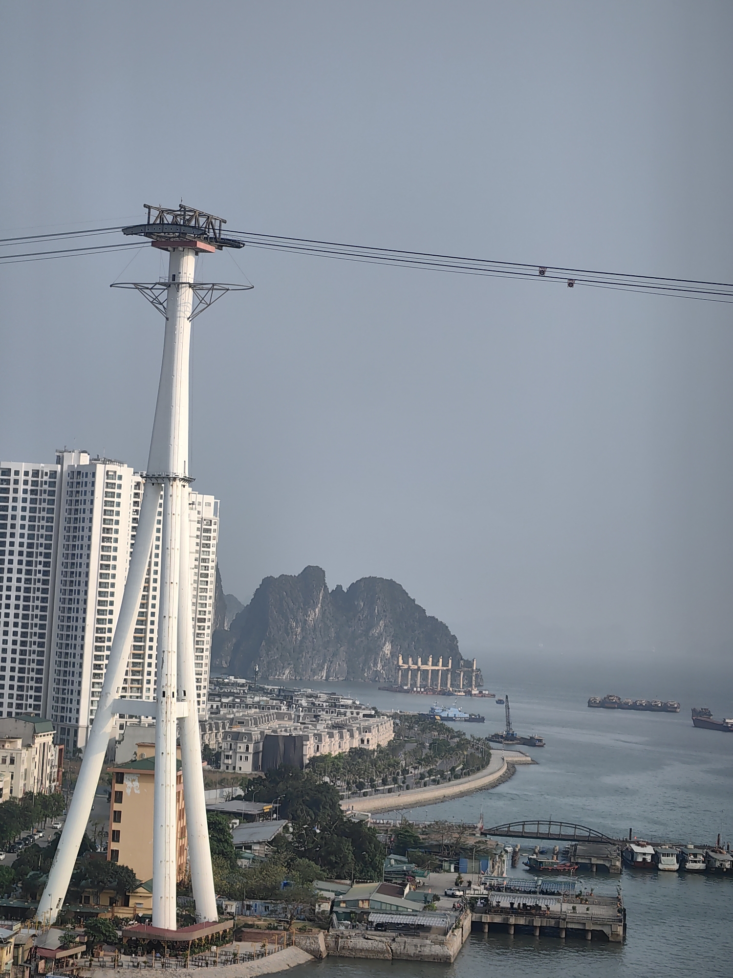 The image shows an aerial view of Bai Chay Ward, Vietnam. A prominent white cable car tower stands in the foreground, with supporting structures reaching the ground. Behind it are several high-rise apartment buildings and other structures. Further back, across the water, is a large island with rocky cliffs. In the water, numerous ships and boats are visible, including a large container ship and several smaller vessels. The shoreline is developed with roads, buildings, and greenery. The sky is overcast, creating a hazy atmosphere.