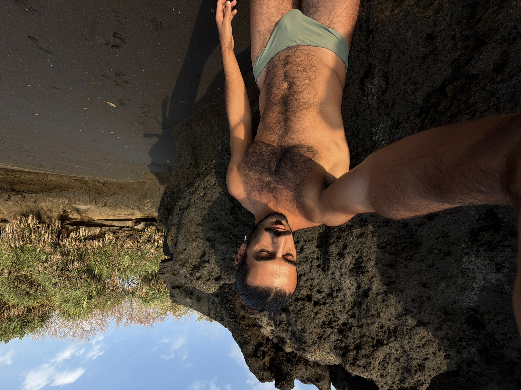 A man with dark hair pulled back into a bun, a mustache, and a goatee stands on a black sand beach in Selemadeg Timur, Indonesia. He is wearing light green swim trunks and is holding a camera to take a selfie. His chest and abdomen are covered in dark hair. Behind him, a large dark volcanic rock formation rises up, partially obscuring the vegetation and sky. The sand is wet in some areas, reflecting the light. The sky is blue with scattered white clouds.