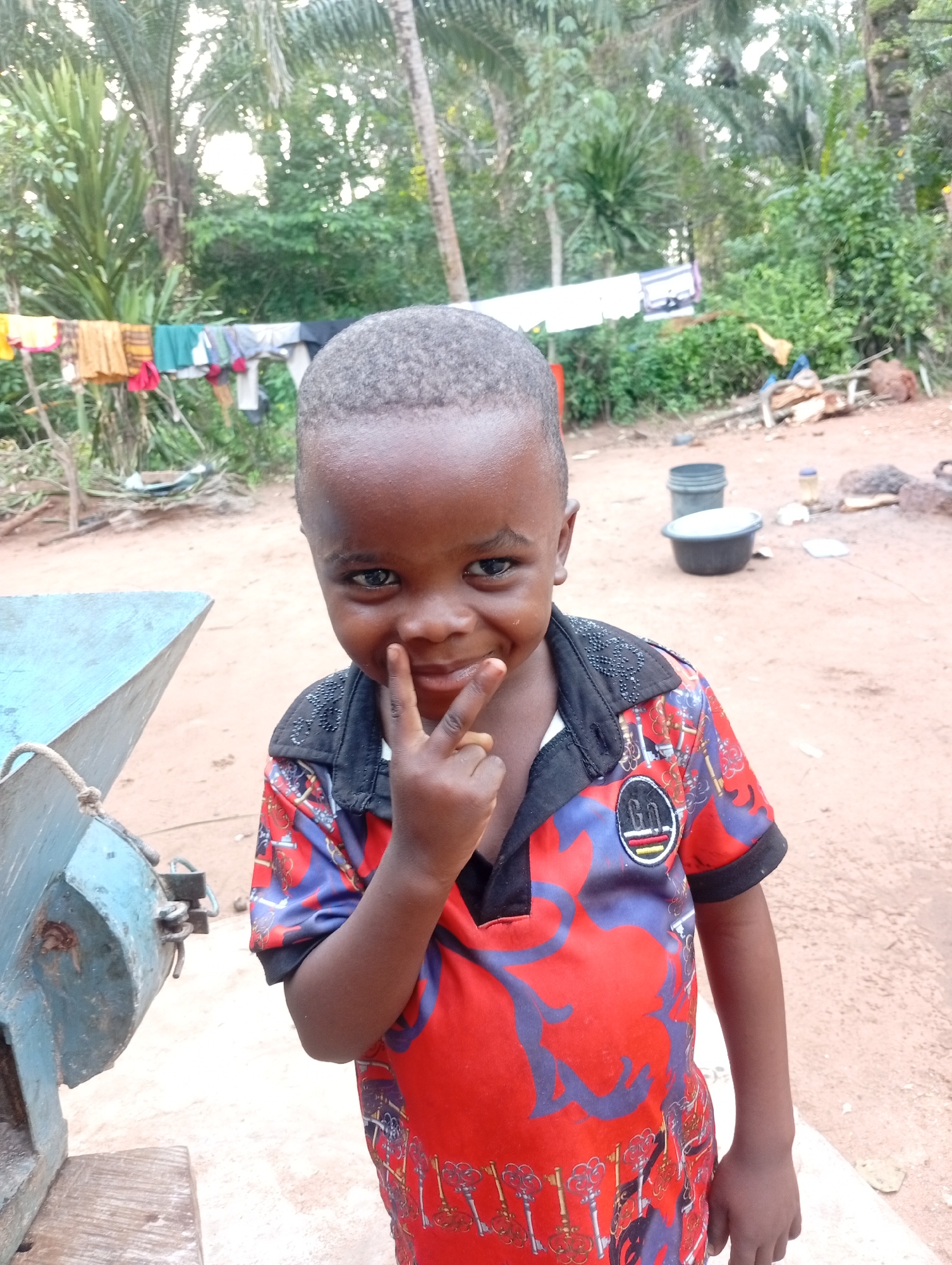 A young Black child, likely a boy, poses for a photo outdoors in a dusty, rural area. He is wearing a red, blue, and purple patterned shirt with a black collar. His left hand is raised to his face, forming a peace sign. In the background, laundry hangs on a clothesline between trees, and various buckets and objects are scattered on the ground. The environment suggests a village setting in Adda, Nigeria, characterized by natural vegetation and simple living conditions. The child's expression is playful, with a slight smile and direct eye contact with the camera.