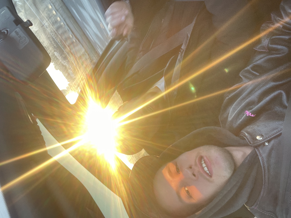 The scene depicts the interior of a vehicle, implied to be located in Caluire-et-Cuire, France. A male individual is prominently featured in the foreground, facing the camera, suggesting a selfie or direct photograph. He wears a dark hooded jacket, potentially leather, with metallic buttons and a pink embroidered insignia on the left chest, and a dark baseball cap beneath the hood. He has short facial hair on his chin and upper lip, and his mouth is slightly open. Intense sunlight streams through the front windshield, creating significant glare and lens flares, directly illuminating the left side of the foreground individual's face. In the background, partially obscured by the solar glare, the right arm and hand of a second individual are visible, gripping an interior element, possibly a door handle or vehicle frame. The strong light indicates bright outdoor conditions, likely sunrise or sunset.