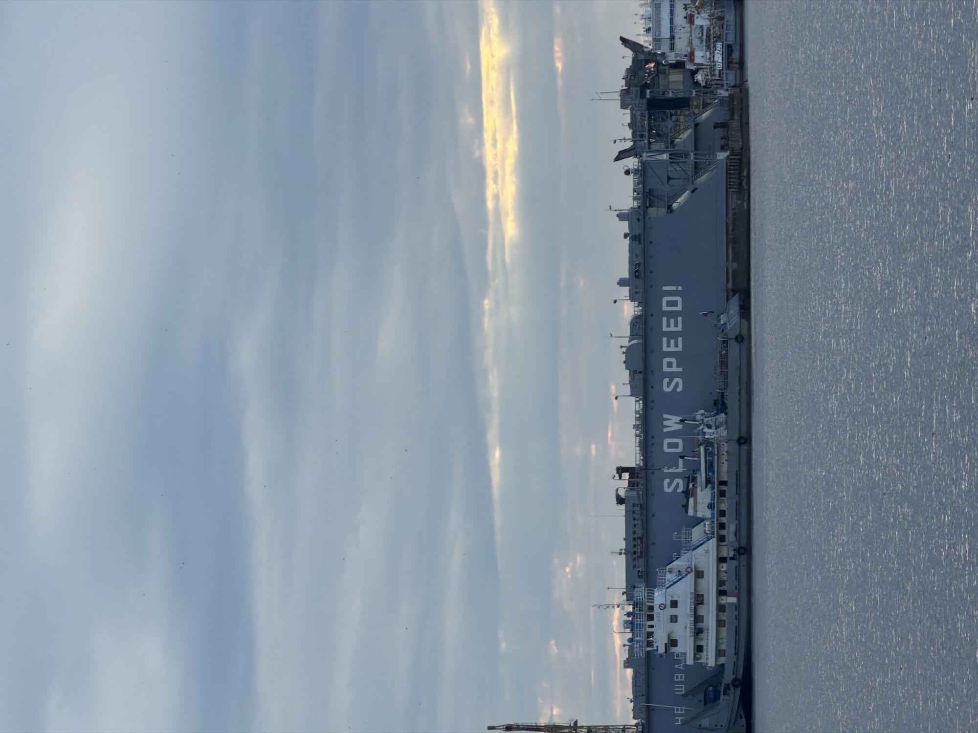 The image depicts a waterfront scene in the city of Chaikovsky, Russia, under a cloudy sky with shafts of sunlight breaking through. In the foreground, a body of water, likely a river or reservoir, exhibits subtle ripples. Dominating the mid-ground are several ships and a large barge. On the barge, the words "SLOW SPEED!" are prominently displayed in large, white capital letters. A white vessel, possibly a tugboat or pilot boat, is docked alongside the barge, its nameplate reading "КАПИТАН ТАРЗУНОВ" (Kapitan Tarzunor). Further to the right, another ship is partially visible, with a number "101-2422" visible on its hull. The background consists of a large, grey structure, possibly a dry dock or a large vessel, with various cranes and industrial elements visible. The overall atmosphere is industrial and maritime, with the cloudy sky suggesting overcast weather. There are no people clearly visible in the image.