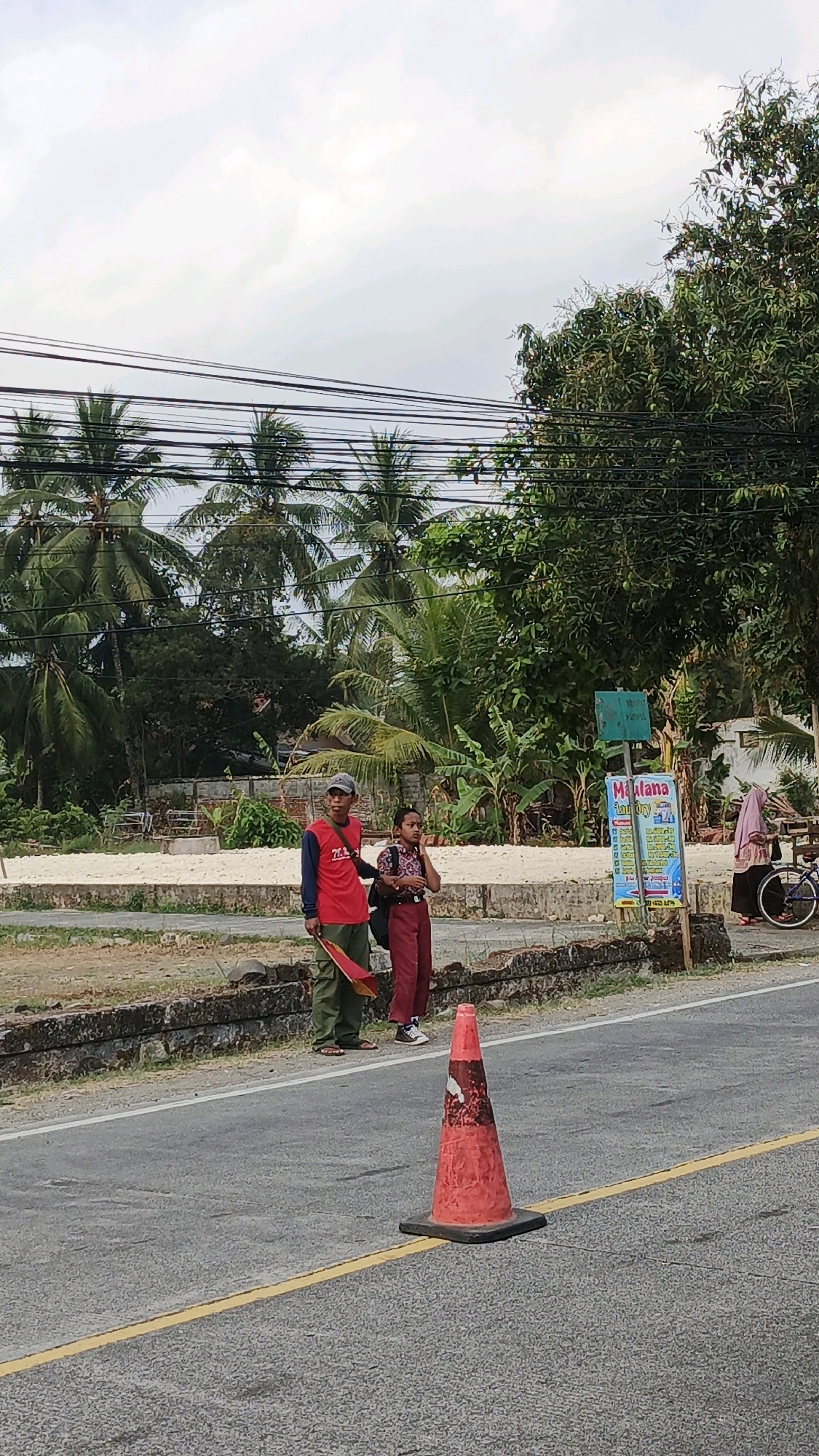 In Cibenda, Indonesia, a street scene unfolds with a road in the foreground, marked by a yellow line and an orange traffic cone. Two individuals stand on the sidewalk, a man in a red shirt and green pants holding a red flag, and a younger person in a school uniform with a backpack. Behind them, a weathered stone wall lines the edge of the road, separating it from a sandy area with sparse vegetation. Lush green trees, including palm trees, dominate the background, with power lines stretching across the frame. A sign advertising "Marlana Laundry" is visible on the right, near a woman in a headscarf and a bicycle. The sky is overcast.