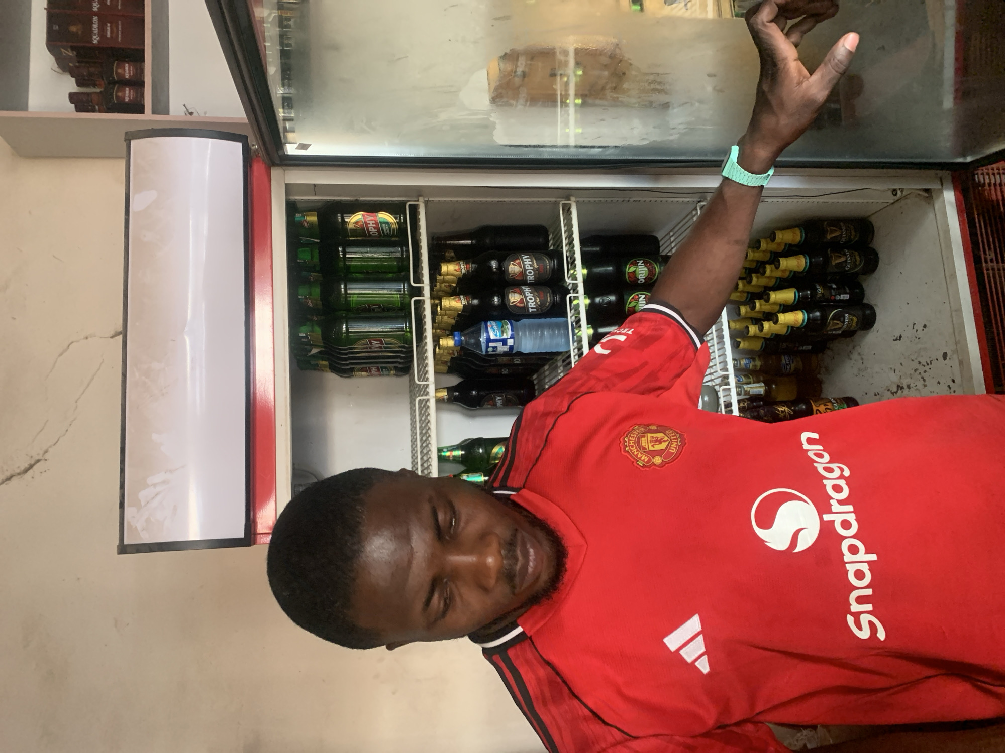 The image shows a man in a red Manchester United soccer jersey standing in front of a refrigerator filled with beverages. The refrigerator contains bottles of Guinness, Trophy beer, and other bottled drinks. The man is partially obstructing the view of the drinks, but they appear to be neatly arranged on shelves within the refrigerator. A wristwatch is visible on his left wrist. The image was likely taken in New Stadium, Nigeria.