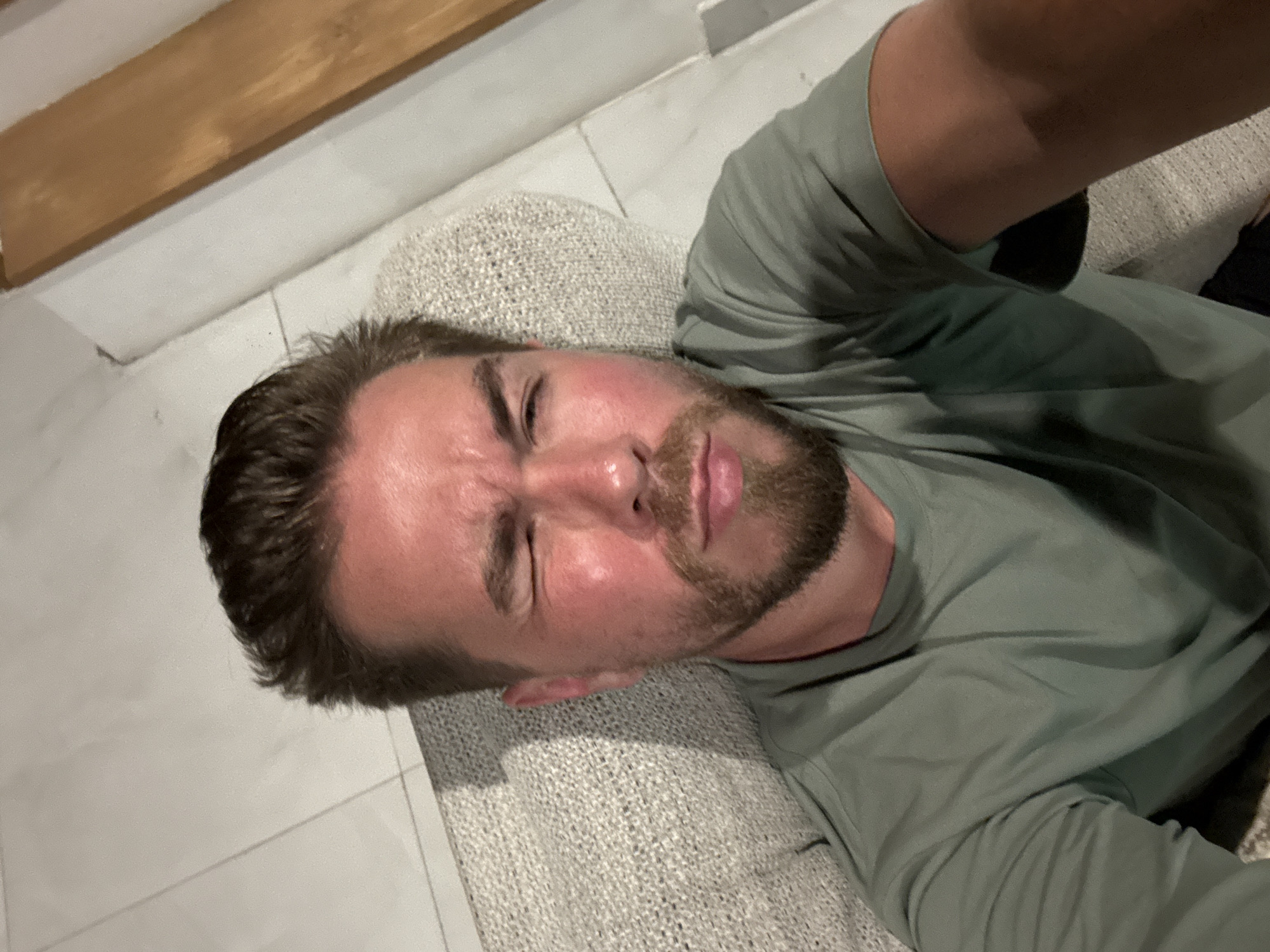 A medium-shot, eye-level photograph taken indoors depicts a man with short brown hair and a beard. He is wearing a light green t-shirt and is lying on a textured light gray surface that could be a couch or cushion. His eyes are partially closed, and his expression is ambiguous. The background consists of a white tile floor and a wooden beam along the top of the frame. Based on the metadata, the location is North Kuta, Indonesia.