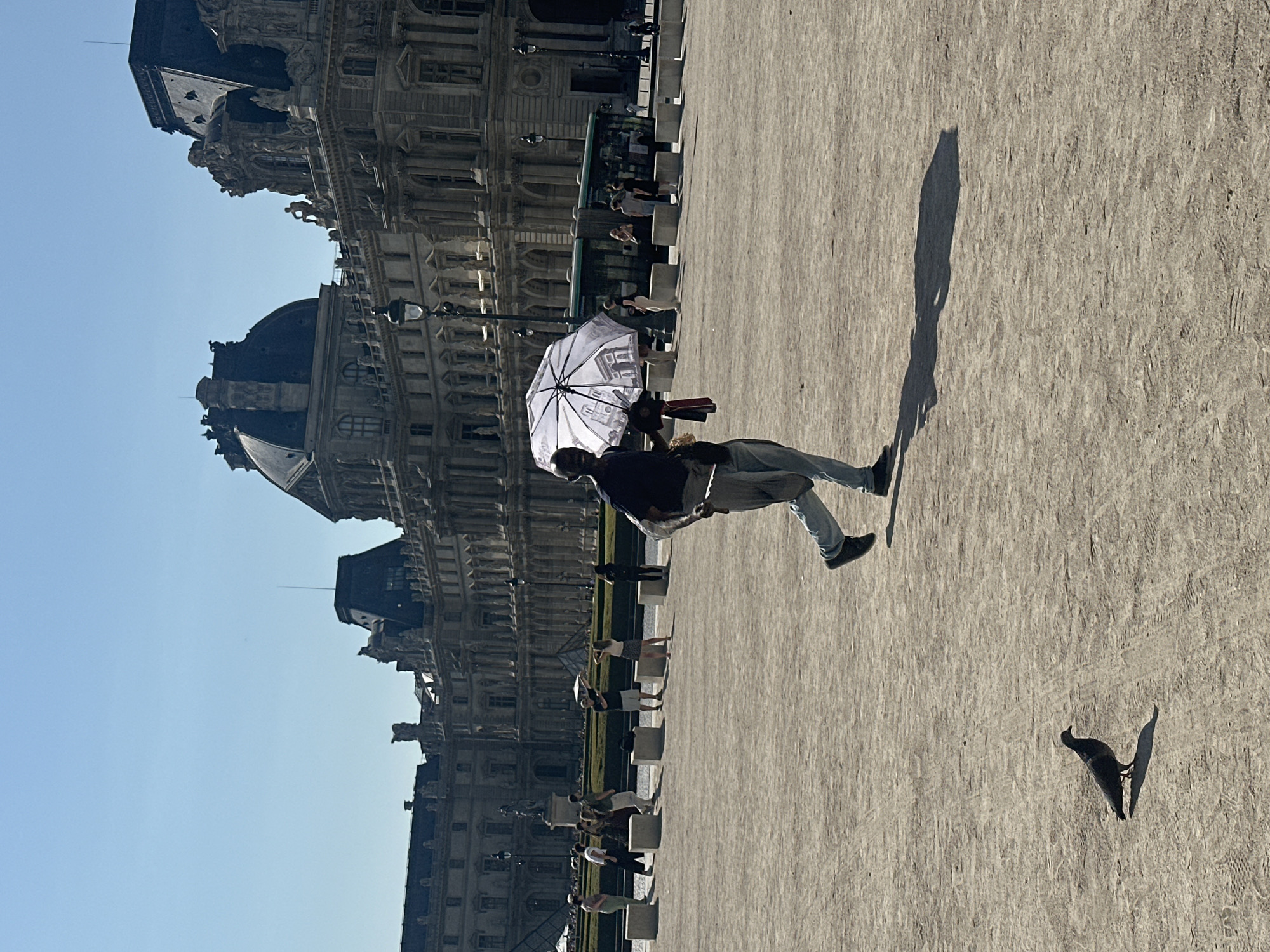 A full, outdoor, eye-level shot captures a bright, sunny day in Paris, France. The scene is dominated by the grand architecture of the Louvre Museum in the background, its ornate facade featuring classical sculptures, mansard roofs, and numerous windows. In the foreground, a dark-skinned man walks from right to left, holding a translucent umbrella above his head. The umbrella is white with intricate black designs, possibly depicting Parisian landmarks. He is dressed in a dark t-shirt, light blue jeans, and dark shoes. He is also carrying a red scarf or cloth in his right hand. To the left of the man, a lone pigeon stands on the dusty ground, casting a shadow. The ground is a light brown, dusty surface with faint tire tracks. In the mid-ground, several people are visible, some walking, others standing, and a green tour bus passes by. The sky is a clear, bright blue.