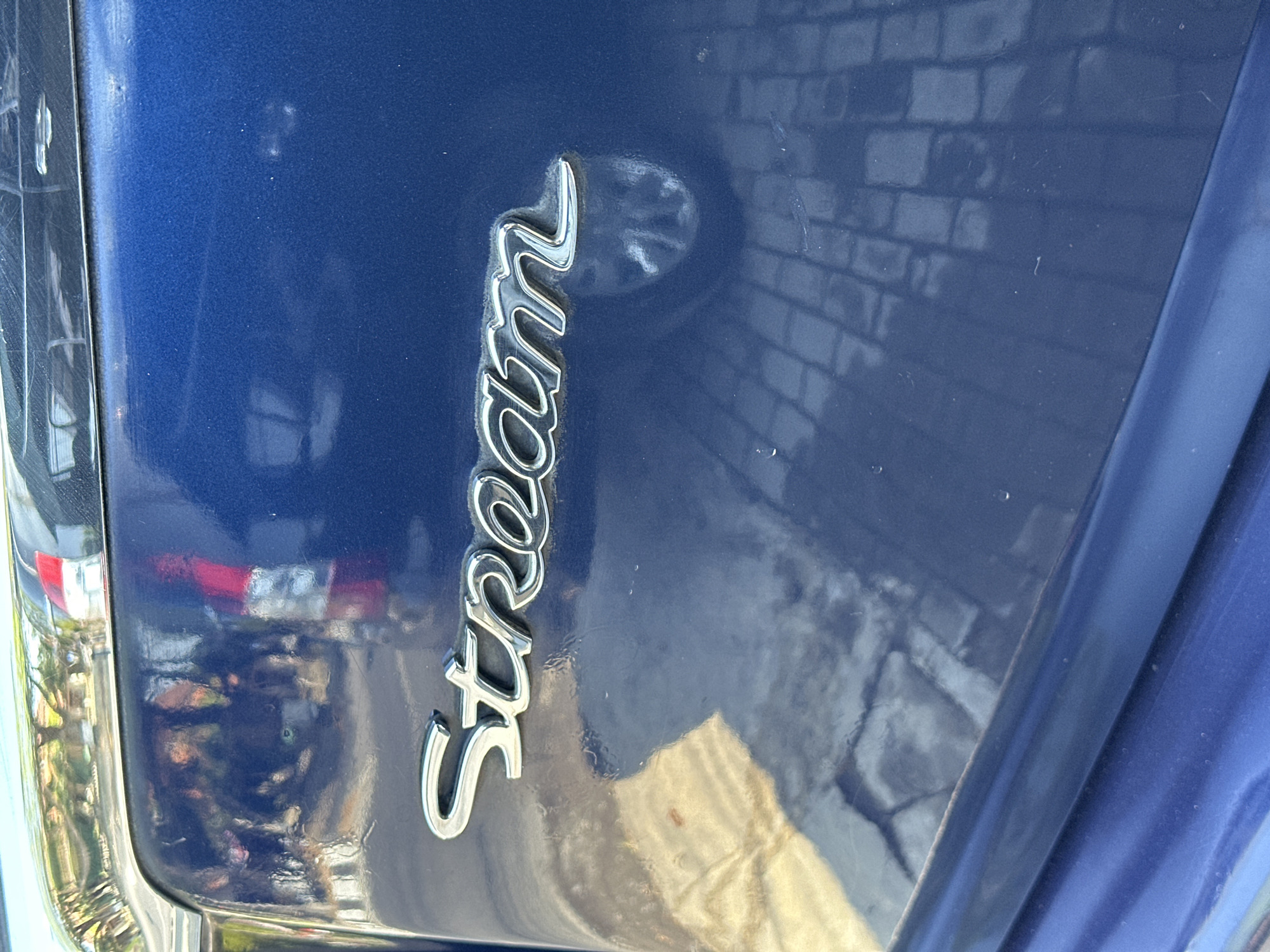 The image shows a close-up of the rear of a dark blue car. A chrome emblem spelling out "Stream" is prominently displayed in the center of the image. Reflections of the surrounding environment are visible on the car's surface, including what appears to be a paved area with a brick pattern and other vehicles. The scene is likely outdoors, given the reflections and lighting. There are no people visible in the frame.
