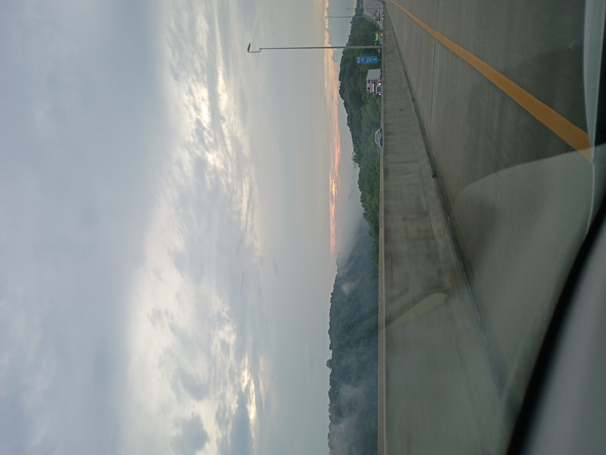 This is an eye-level, medium shot of a highway overpass in Lexington, United States of America. The sky is overcast with a few patches of lighter clouds that appear to be illuminated by sunlight breaking through. A misty fog blankets the tops of a dense forest that extends across the horizon, with a sliver of pale orange visible on the far right, suggesting a sunset or sunrise. The highway has multiple lanes, separated by a solid yellow line on the right side of the frame. A concrete barrier runs along the left side of the overpass, with several vehicles, including trucks and cars, traveling in the same direction. A lone streetlamp stands tall on the right side of the highway. The image is slightly blurry, suggesting it was taken from a moving vehicle.