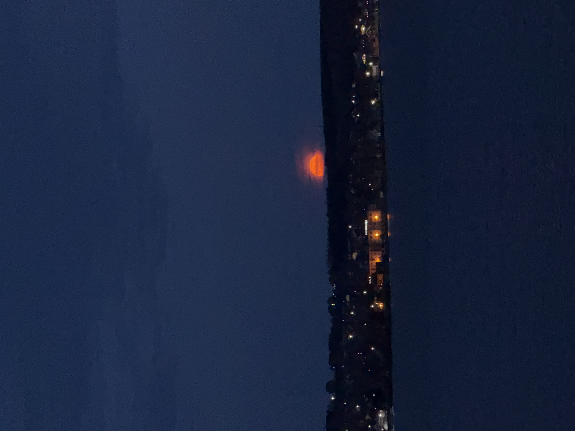 At night, the image shows a partial orange glow emerging between two dark blue expanses. To the right of the orange glow, a darker area appears to contain structures with scattered points of light. The structures and lights could be part of Votkinsk, Russia. There are no people visible in the image.