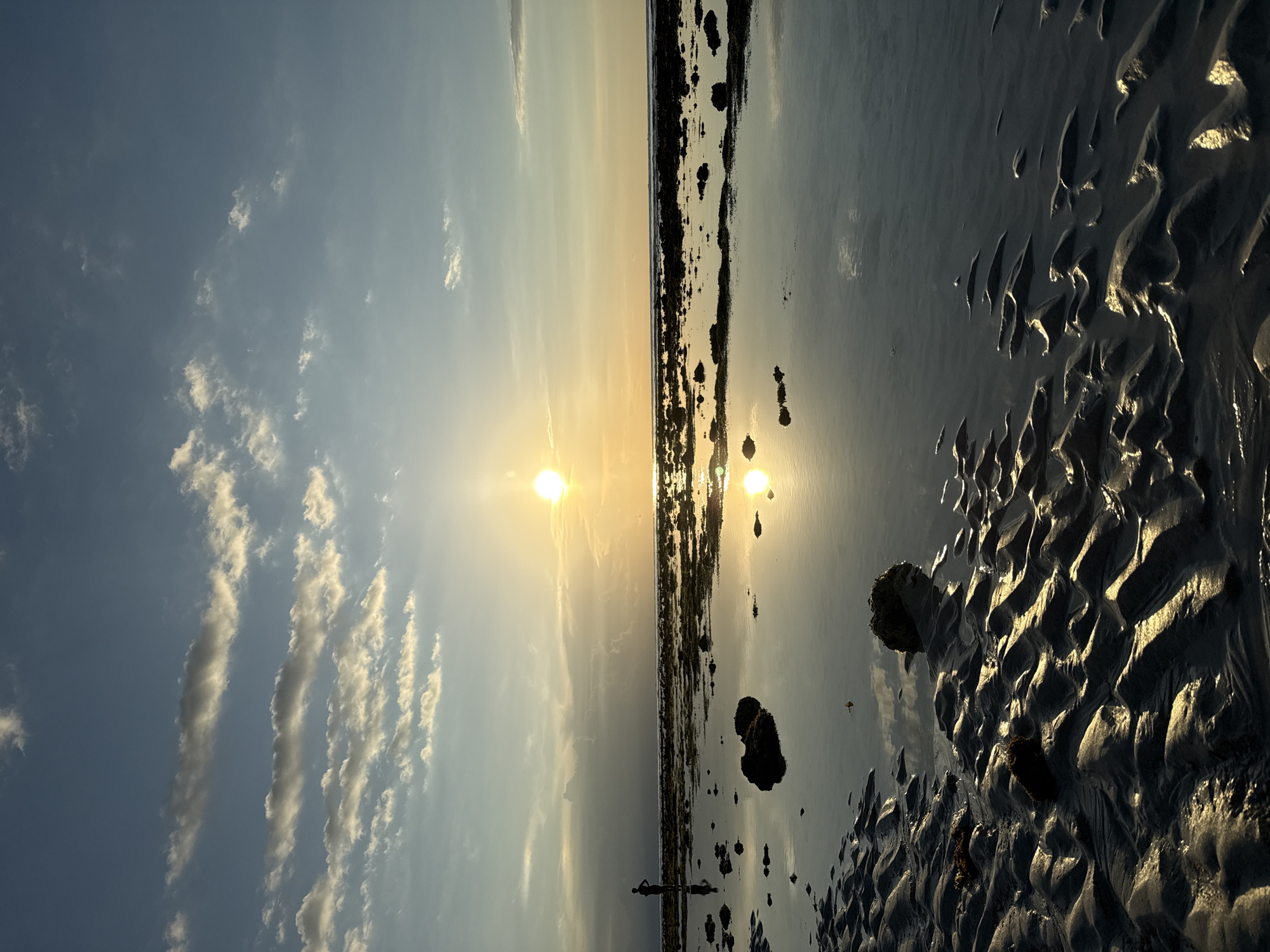 The image captures a coastal scene in Unknown, Indonesia, at either sunrise or sunset, suggested by the low angle of the sun. The sky is a mix of blue and white clouds. A strip of land separates the sky and the ocean. The beach has wet sand patterns. Several rocks of varying sizes are scattered on the beach. The sunlight casts a reflection on the water. There is a figure standing at a distance.