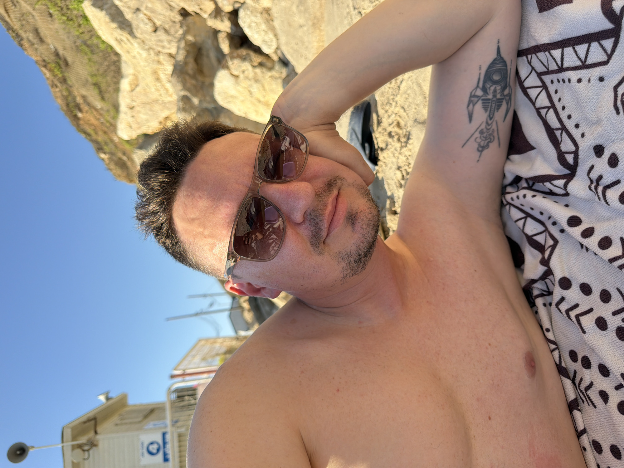 A shirtless man with dark hair and a goatee is lying on a beach towel on a sandy beach. He is wearing sunglasses and resting his head on his hand. His left arm, which is visible, has a tattoo of a rocket. In the background, there is a lifeguard station, a cliff face, and a clear blue sky. This scene is taking place in Bat Yam, Israel, as indicated by the lifeguard station signage.