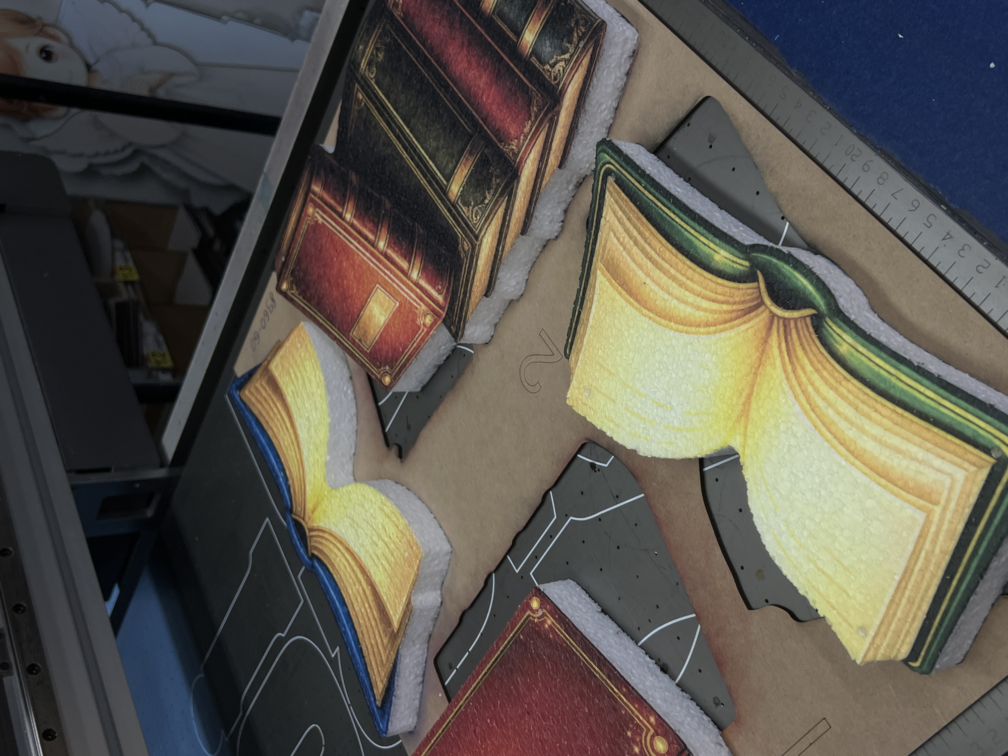 The image captures a close-up, high-angle view of several die-cut shapes resembling books, arranged on a light brown work surface. These shapes feature detailed illustrations; some depict closed books with ornate spines in red, green, and black with gold accents, while others show open books with fanned yellow and white pages and colored bindings (green, blue). The edges of these cutouts have a distinct white, foamy texture, suggesting they were cut from a thicker material like foam board.
Irregular grey pieces, possibly waste material from the cutting process, are interspersed among the book shapes. On the right side of the frame, a metal ruler or cutting guide with metric measurements (centimeters) is visible. A faint, reversed numeral "2" is subtly marked on the brown surface.
In the upper left portion of the image, a dark, reflective surface shows an upside-down reflection of what appears to be a person or a doll with light hair and a white garment. A metallic track or rail structure is visible along the left edge, hinting at industrial machinery such as a large-format printer or a cutting device. The scene suggests an indoor workshop or production environment. Faint, upside-down text "99-0968" is also discernible within the reflection.