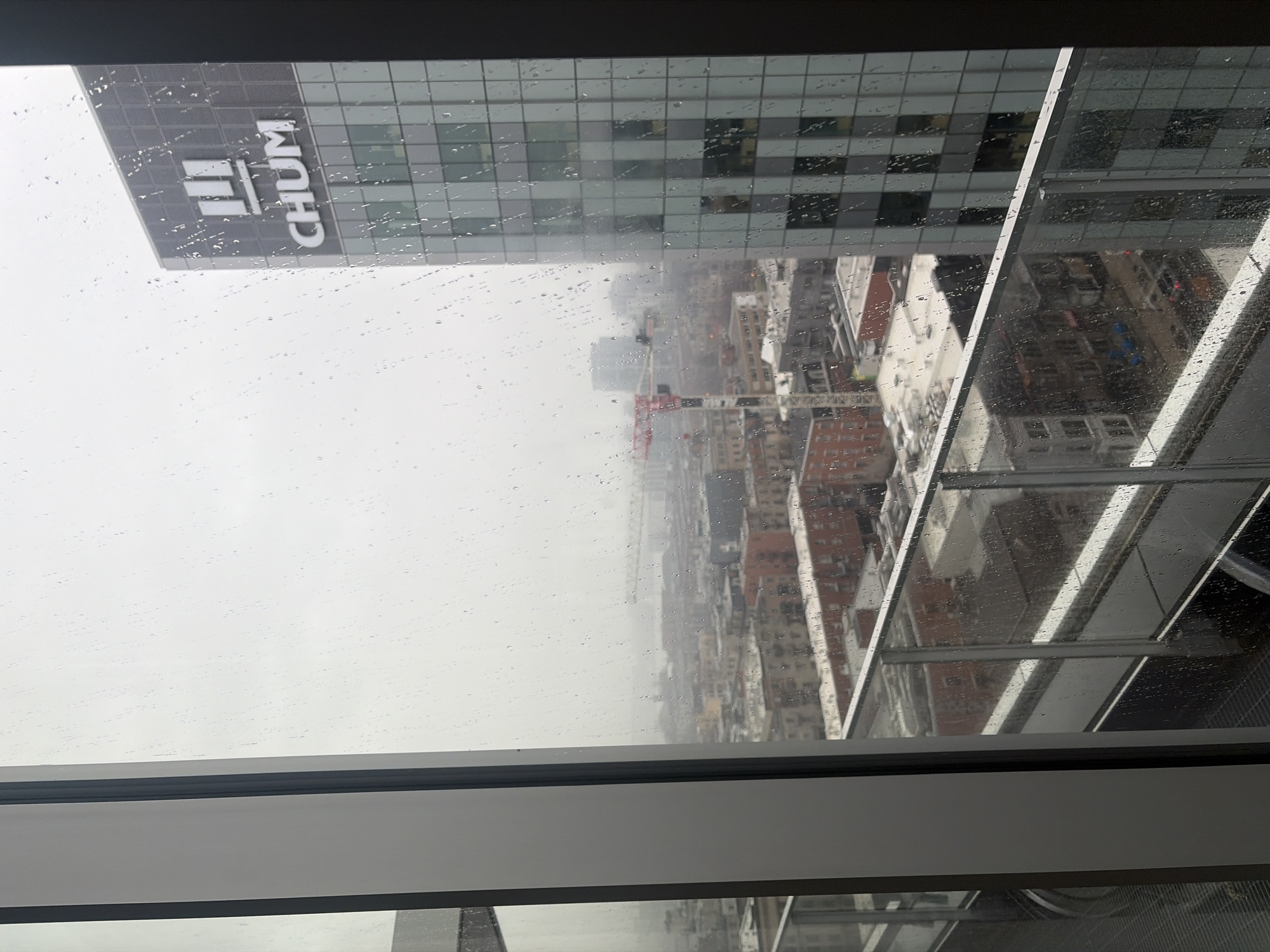 The image, captured from a high-rise window, presents an overcast and rainy day over a dense urban landscape, likely Montreal, Canada. The windowpane in the immediate foreground is covered in countless small water droplets, indicating precipitation.
Through the rain-speckled glass, a prominent modern building with a grey glass facade stands to the left. Its side clearly displays the white, capitalized text "CHUM" accompanied by a logo consisting of three vertical bars, identifying the Centre hospitalier de l'Université de Montréal.
Beyond this landmark, a construction site is visible in the mid-ground, featuring a tall red and white crane. The city stretches into the distance under a uniform, bright grey sky, filled with a variety of buildings whose details are somewhat softened by the atmospheric conditions. Another reflective building or structure is partially seen on the right side, also wet from the rain. The overall scene conveys the bustling activity of a city experiencing gloomy, wet weather conditions during the daytime.