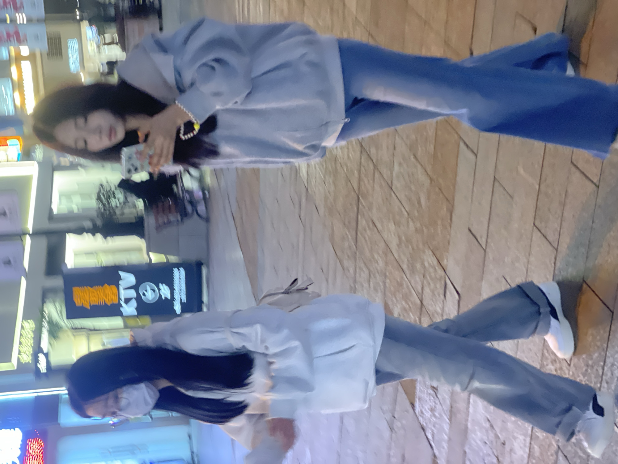 The scene depicts two young adult females walking on a paved urban sidewalk at night within the Chongqing High-tech Zone, China. The ground appears to be composed of rectangular tiles, with some reflective areas suggesting dampness or wetness.
The individual on the left is wearing a white face mask, glasses, a light grey hooded sweatshirt, light blue wide-leg jeans, and white sneakers with black accents. Her long, dark hair is visible. She carries a light-colored backpack over her right shoulder and is looking generally forward and slightly left.
The individual on the right, walking slightly behind and to the right of the first person, has long, dark hair. She is wearing a light blue hooded sweatshirt, dark blue wide-leg pants, and partially visible white shoes. Both hands are holding a light-colored smartphone with a patterned case, and her gaze is directed at the screen. A white beaded bracelet with a yellow charm is visible on her left wrist.
The background is a blurred nighttime urban environment, illuminated by various artificial lights. Visible elements include a vertical sign with "KTV" and Chinese characters in orange and white, other glowing building facades, and neon signs, indicating a commercial or entertainment district. Both individuals are in motion, walking from left to right across the frame.
