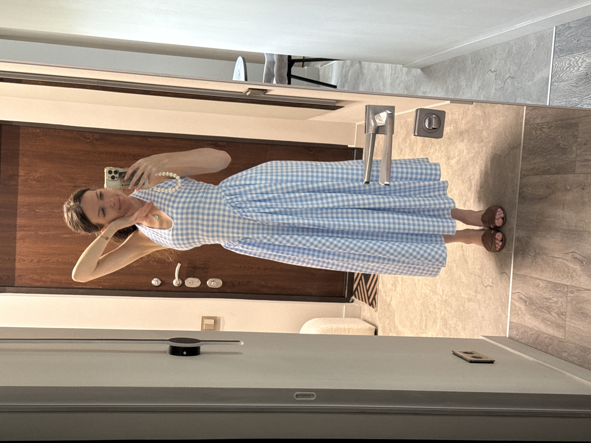 A young woman is captured in a mirror selfie, standing upright and looking towards the camera with a soft smile. She appears to be in her 20s or 30s, with light brown hair. She is wearing a light blue and white checkered (gingham) sleeveless dress, a pearl necklace, a bracelet, and a ring on her left hand. Her right hand holds a smartphone, likely an iPhone, in front of her face to take the picture, while her left hand is bent near her ear. She is wearing brown sandals.
The setting is an indoor space, possibly an apartment or hotel room in Dolgoprudny, Russia. Reflected in the mirror behind her is a dark brown wooden door with metal handles and a lock. To the right of the door, a wall with light-colored, textured tiles is visible, and the floor features a mix of light-colored and wood-patterned tiles. In the upper reflection, hints of furniture like a table and chairs can be seen.
The mirror is framed by a light-colored surface, possibly a vanity or wall panel. On this surface, a small black circular device with a silver accent and a rectangular wooden object with two holes are visible. A power outlet is also present on the right edge of the mirror's frame. The lighting suggests it is daytime. No discernible text is visible on clothing or objects within the reflection.
