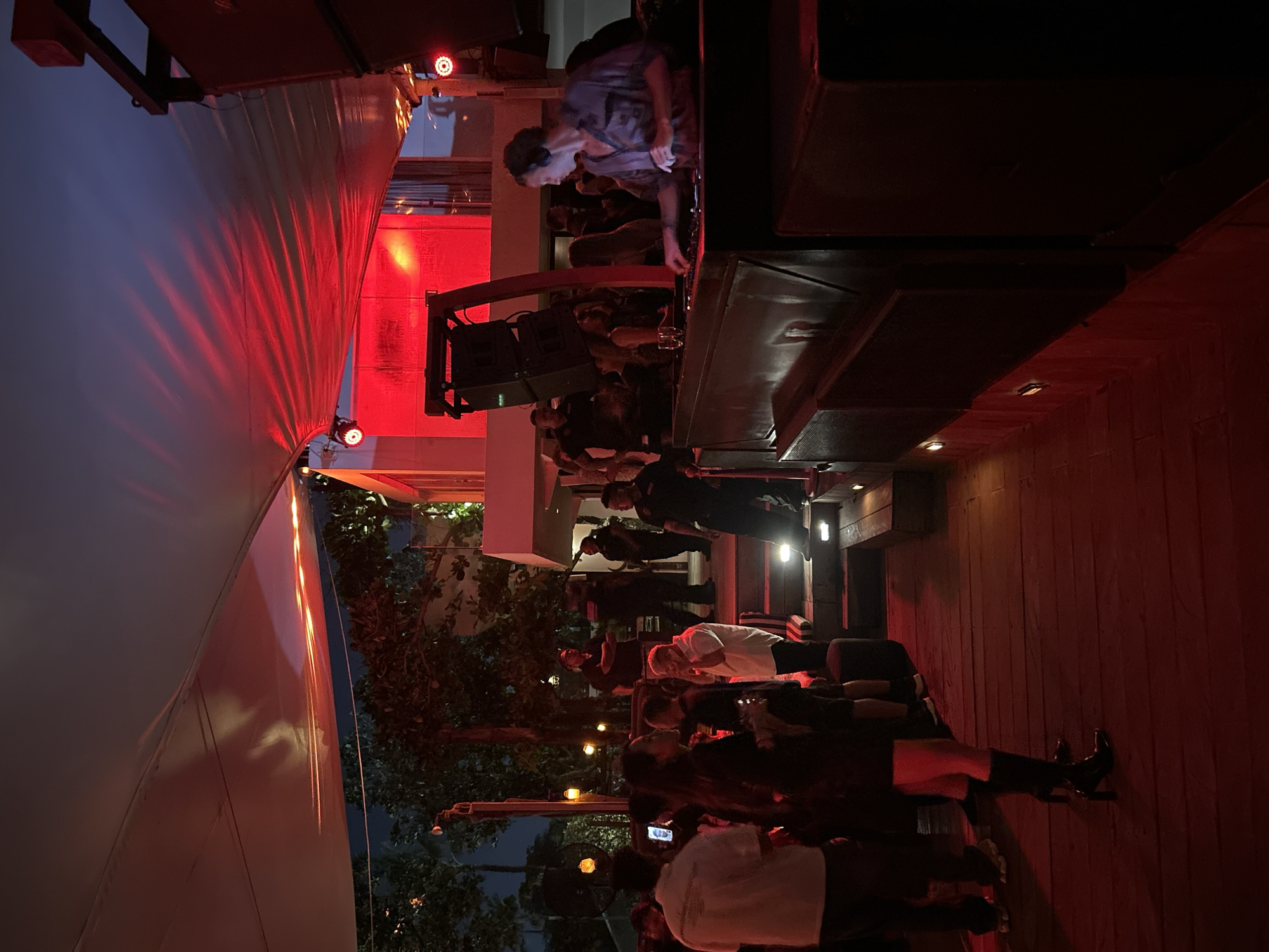 The image depicts an outdoor nighttime gathering at a venue in North Kuta, Indonesia, characterized by red lighting and a festive atmosphere. In the foreground, a woman wearing black boots and a black dress stands on a wooden platform, facing away from the camera. To her right, a DJ with headphones is actively working at a console, with a crowd visible behind him. The DJ booth features large speakers and a red-lit structure. The background reveals a building with an awning illuminated by red lights, casting patterns on the fabric. Several individuals are scattered throughout the scene, some appearing to be security personnel. Trees and foliage are visible in the periphery, suggesting an outdoor or semi-outdoor setting. The overall ambiance suggests a lively event, possibly a party or a club night.