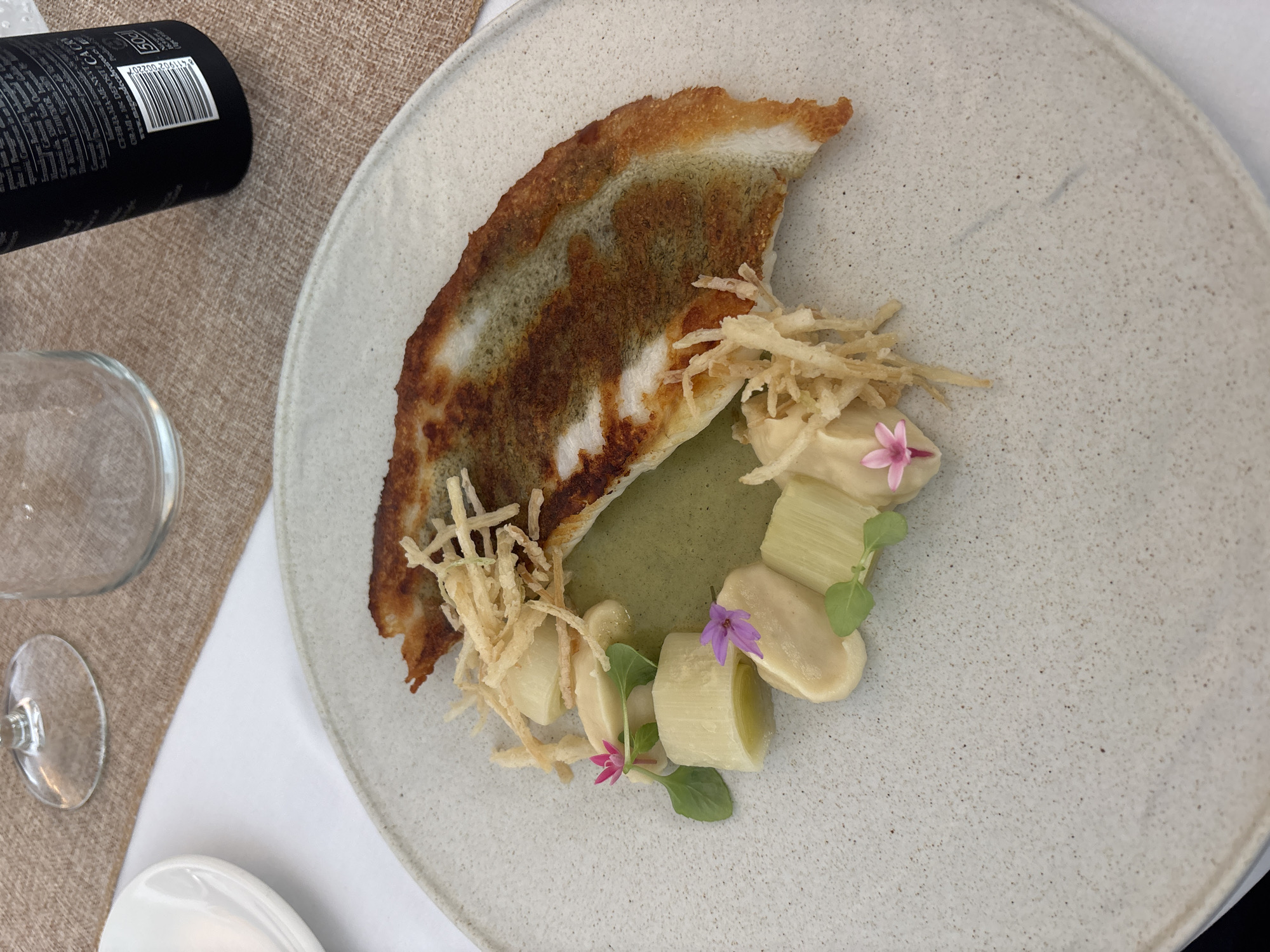 A single plate of food is presented on a textured placemat. The plate is round and light gray with dark speckles. On the plate, there is a piece of pan-fried fish with crispy skin, resting in a pool of light green sauce. Arranged around the fish are several pieces of white leek, some dollops of a creamy white sauce, and a garnish of fried potato strings. Two small pink flowers and a few green leaves are placed as decorative elements. In the background, a wine glass and another glass are visible, along with a white side plate. The setting appears to be a restaurant in Carboneras, Spain.