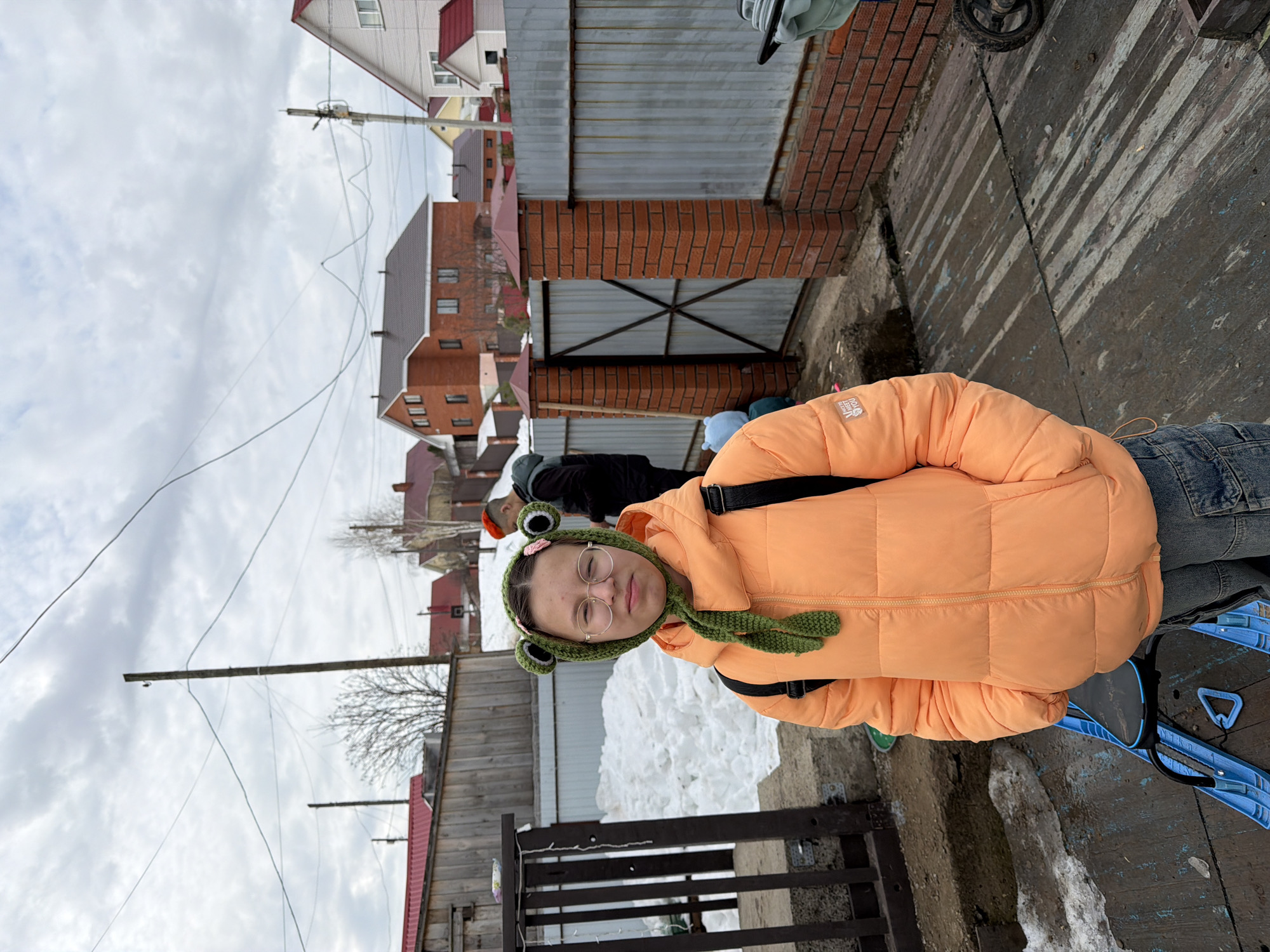 A young person with glasses and a frog-themed hat is standing in front of a fence with snow piled behind it, wearing an orange puffer jacket. Behind the fence, there are houses and a cloudy sky. The person has a blue sled. Overhead power lines are visible. The location is likely Chaikovsky, Russia.