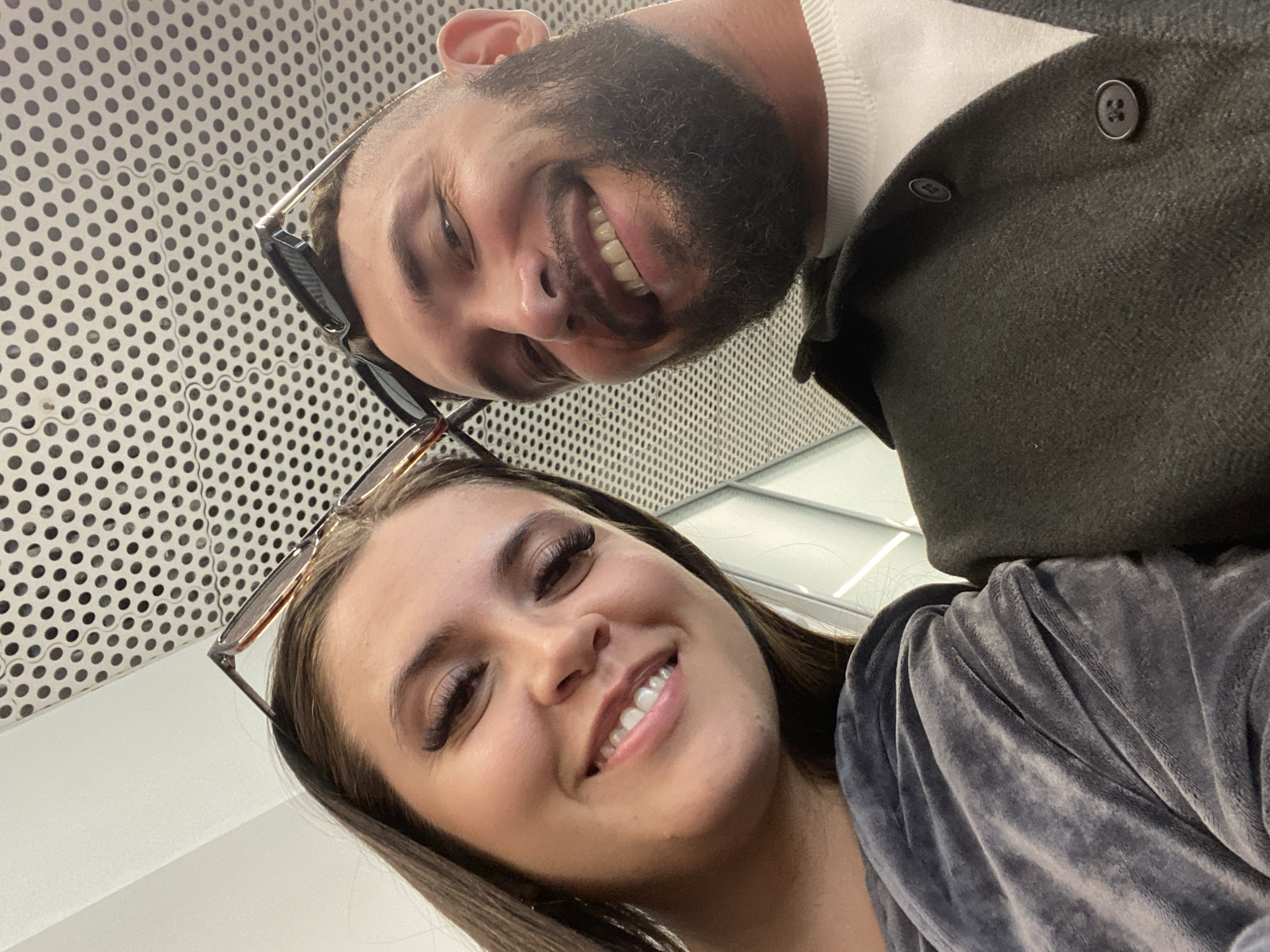 Two people are captured in a close-up selfie. The person on the left, appearing to be a woman, has long brown hair and is smiling, her head tilted slightly. She is wearing a grey velvet top. The person on the right, a man with a beard, is also smiling. He is wearing a dark jacket over a white sweater. Both individuals are wearing sunglasses perched on their heads. The background features a perforated ceiling. The specific location in Новое Мартьяново, Russia is not discernible from the image.