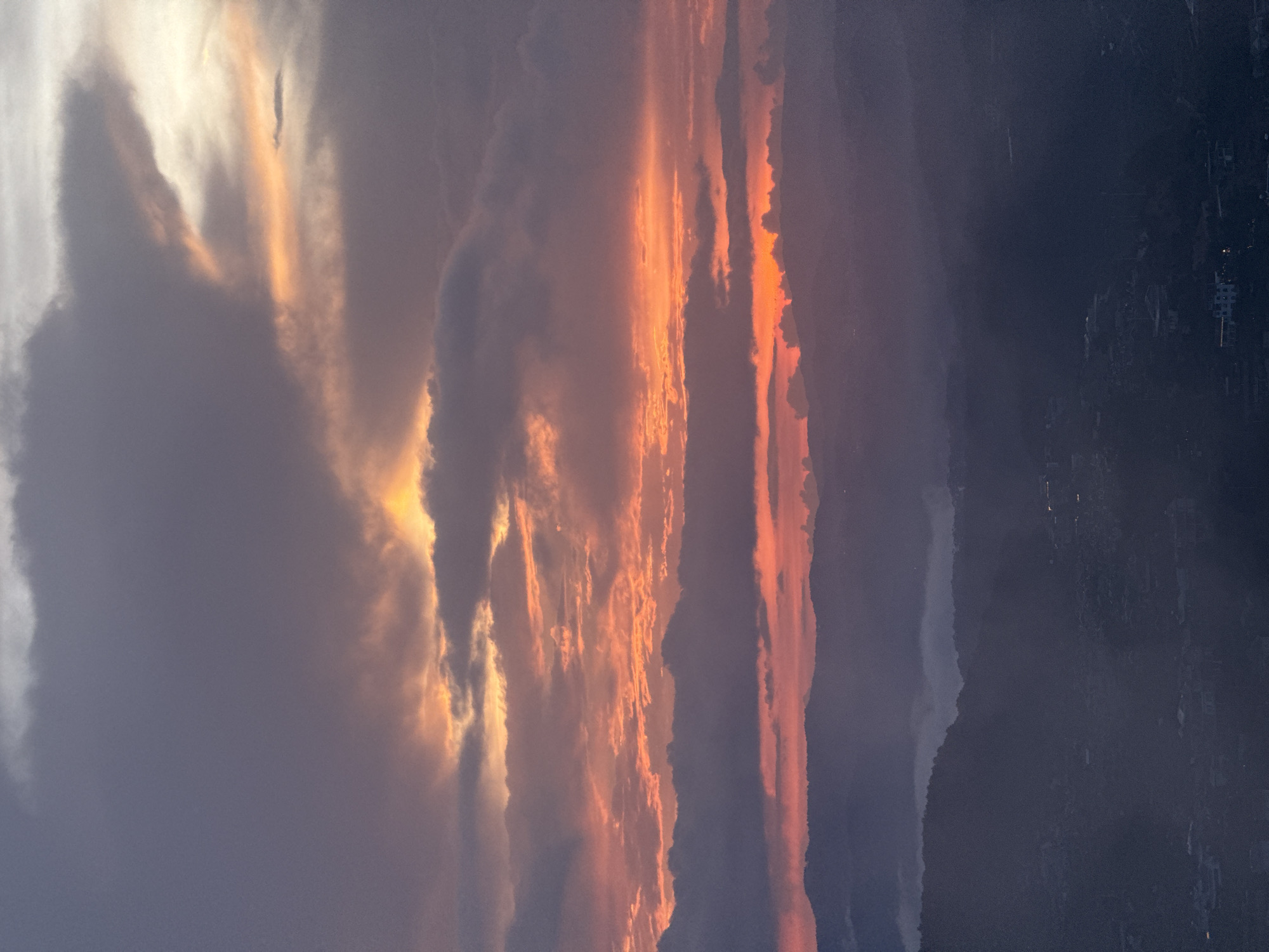 The image depicts a sunset sky over Trường Thọ, Vietnam. A combination of dark, heavy clouds and vibrant orange and yellow hues dominates the scene. In the distance, faint lights suggest the presence of a cityscape. The darker foreground appears to be a mountainous or hilly landscape.