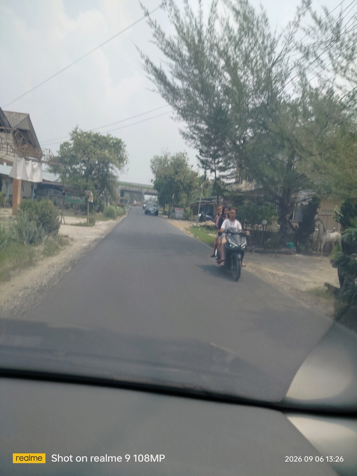 A street scene in Melatikebun, Indonesia, shows a paved road with a motorcycle carrying three individuals, two women and a person driving, traveling away from the viewer. A white horse statue stands on the right side of the road. Buildings and trees line the street. The sky is overcast. A watermark in the lower left corner reads "realme Shot on realme 9 108MP" and in the lower right corner "2026 09 06 13:26".