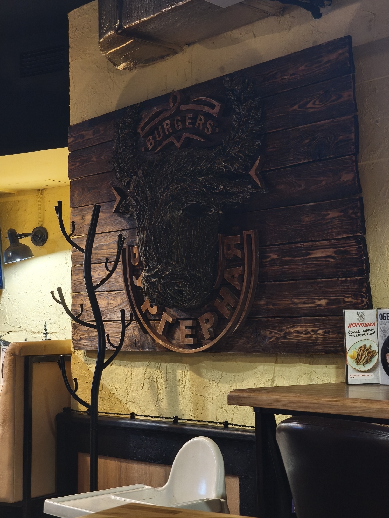 The image captures the interior of a restaurant or cafe, characterized by a rustic and warm aesthetic. The central focus is a large, dark wooden sign mounted on a textured, yellowish wall. The sign features a detailed, stylized relief carving of an animal head, possibly a bull or boar, with "BURGERS." inscribed above it and "БУРГЕРНАЯ" (Burger Joint) below in a semi-circular wooden frame.
In the foreground and to the left of the sign stands a black, tree-branch-like metal coat rack. Further left, a wall-mounted industrial-style lamp casts a warm glow on the textured wall and a tan upholstered booth. To the right of the wooden sign, a dark wooden table with a matching dark chair is visible, positioned in front of another upholstered seating area. A white high chair with a tray is partially visible in the bottom-center foreground, suggesting a family-friendly establishment.
Two menu cards or promotional brochures are visible on the table to the right. One prominently features "КОРЮШКА" (Smelt), displaying an image of fried fish and the text "СОЧНАЯ, РУМЯНАЯ, ХРУСТЯЩАЯ, ТВОЯ!" (Juicy, ruddy, crispy, yours!) along with "ЖАРЕНАЯ КОРЮШКА 690₽" (Fried Smelt 690 rubles). The other card's top header reads "ОБЕД" (Lunch).
The overall ambiance is cozy and inviting, illuminated by warm artificial light. No people are present in the scene. The Cyrillic text and ruble currency indicate the location is in Russia.