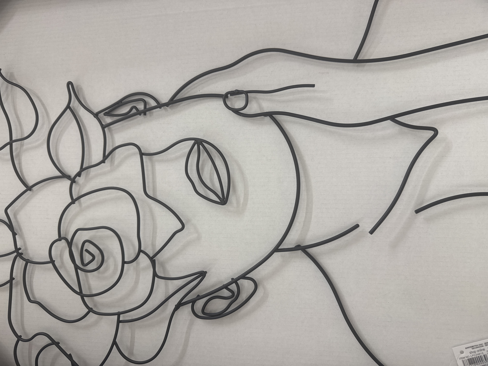 This is a close-up, high-angle shot of a black wire art sculpture of a woman's head and upper body adorned with a rose. The sculpture is mounted on a light-colored, corrugated cardboard background. The art depicts a profile of a woman with her hand raised to her cheek, surrounded by abstract lines forming a rose. The sculpture is minimalist, using a single continuous line to create the forms. There is a small white label attached to the bottom left corner of the cardboard, which appears to be a product tag with text and a barcode. The lighting is even, casting soft shadows. The sculpture does not appear to be located in Kato Polemidia Municipality, Cyprus.