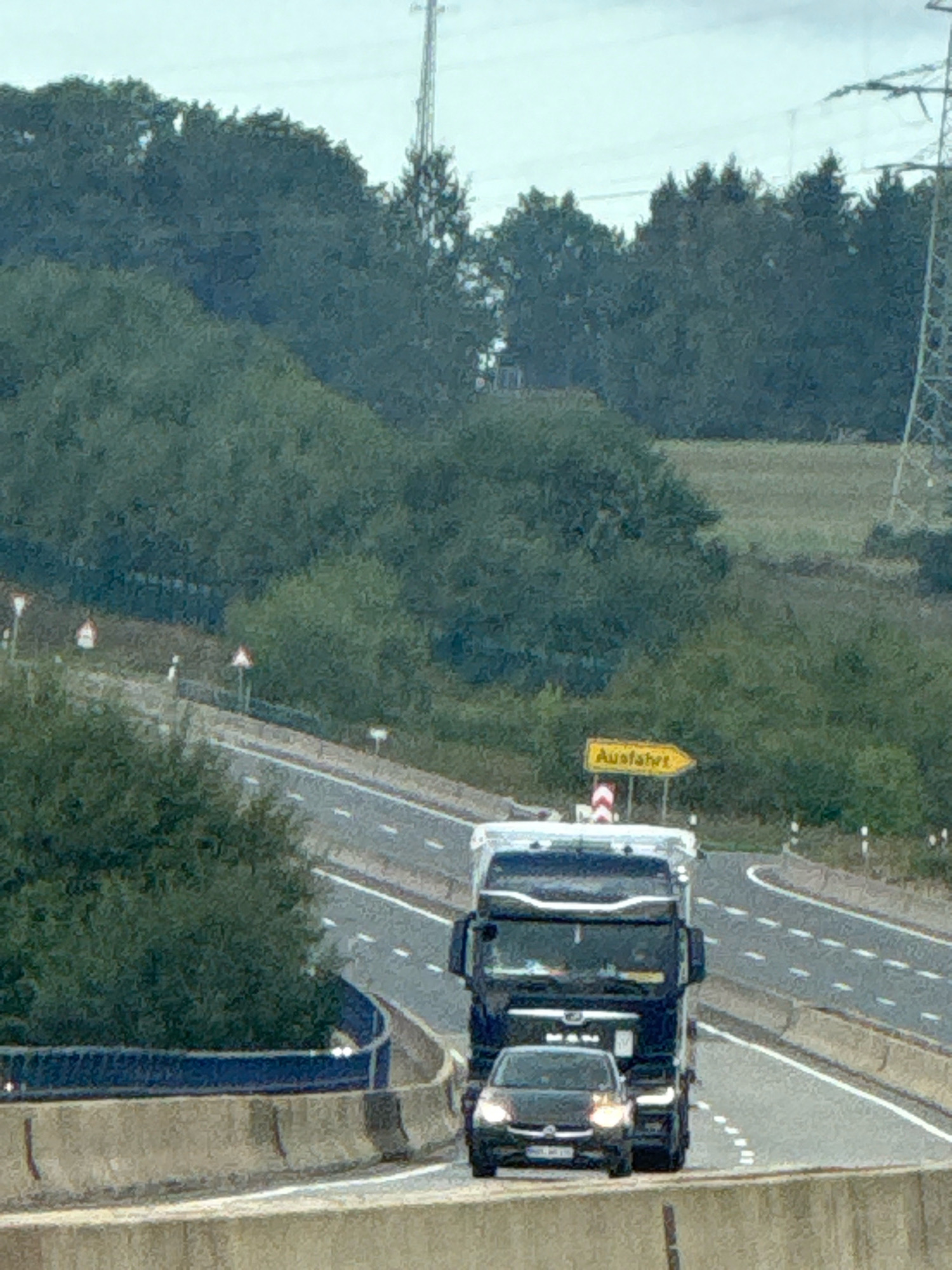 The image shows a view from an overpass looking down at a multi-lane highway in or near Bad Lauterberg im Harz, Germany. A dark-colored passenger car is traveling behind a large, dark blue semi-truck on the highway. A yellow directional sign with the German word "Ausfahrt" (meaning exit) is visible to the right of the truck. The highway is flanked by concrete barriers and surrounded by dense green trees and foliage, suggesting a rural or semi-rural setting. A power line tower is visible in the background. The sky is overcast.