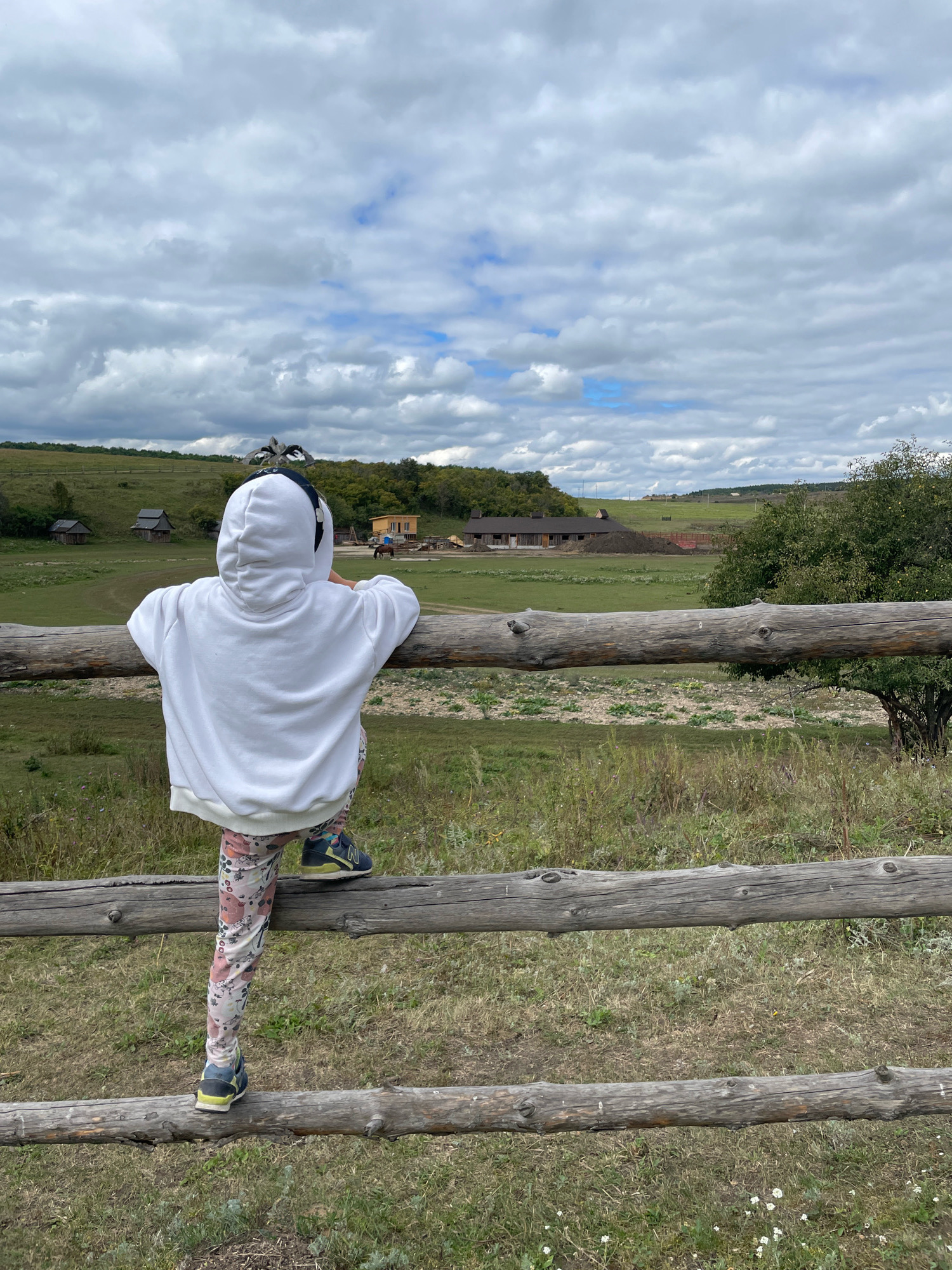 A child, viewed from behind, stands on a wooden fence overlooking a rural landscape. The child is wearing a white hooded sweatshirt and patterned leggings, with one foot on the upper fence rail and the other on a lower rail. The child's head is turned slightly to the left, and they are wearing headphones. The fence is made of rough-hewn logs. In the background, there are rolling green hills, several small wooden buildings, and a larger barn-like structure. The sky is filled with dramatic, cloudy formations, with patches of blue sky visible. The overall impression is one of a serene, pastoral setting on an overcast day.